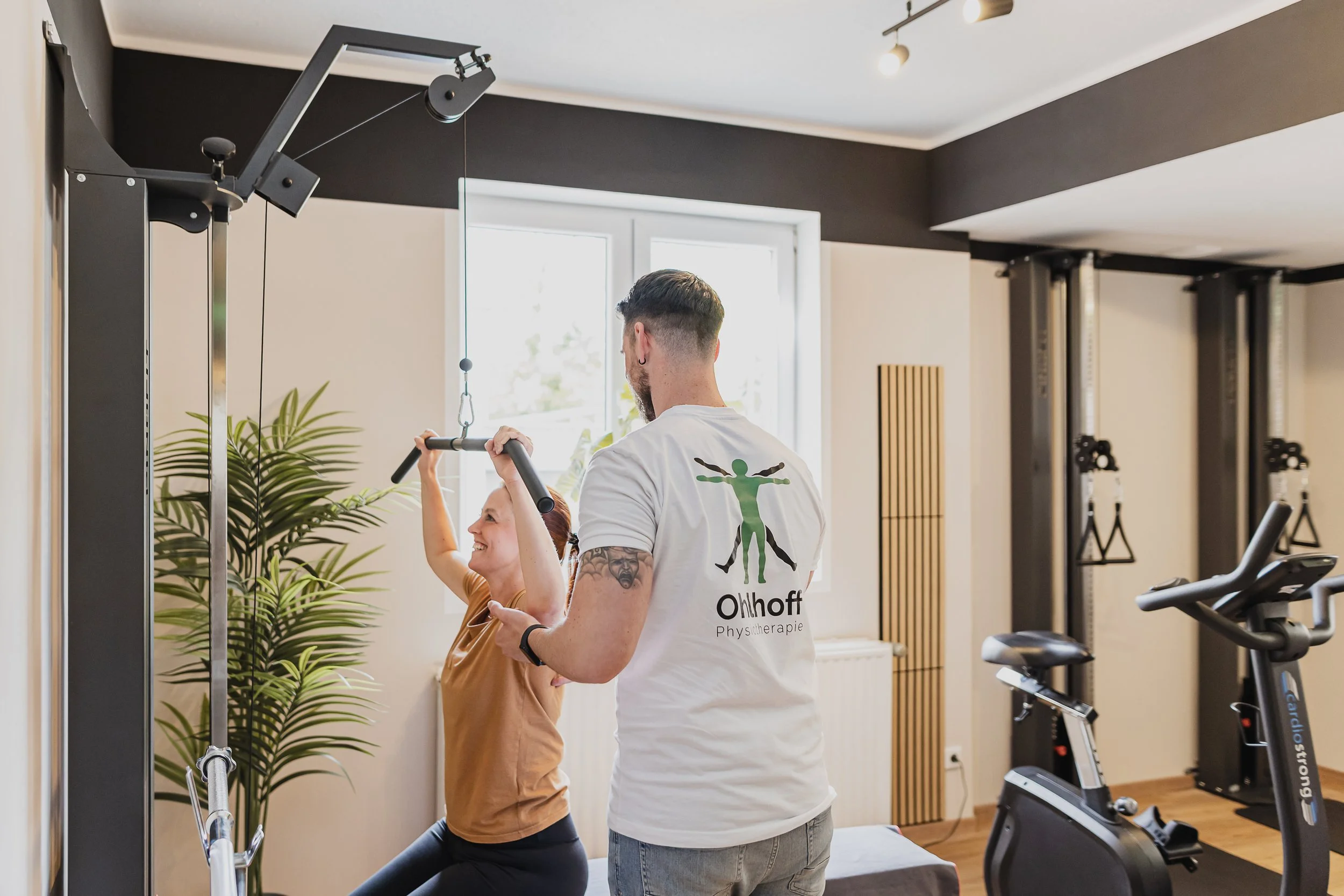 Ein Physiotherapeut führt mit einer Patientin eine Übung mit einem Seil an einem Kraftgerät aus. Im Raum stehen weitere Trainingsgeräte, und es gibt ein Fenster mit natürlichem Licht.