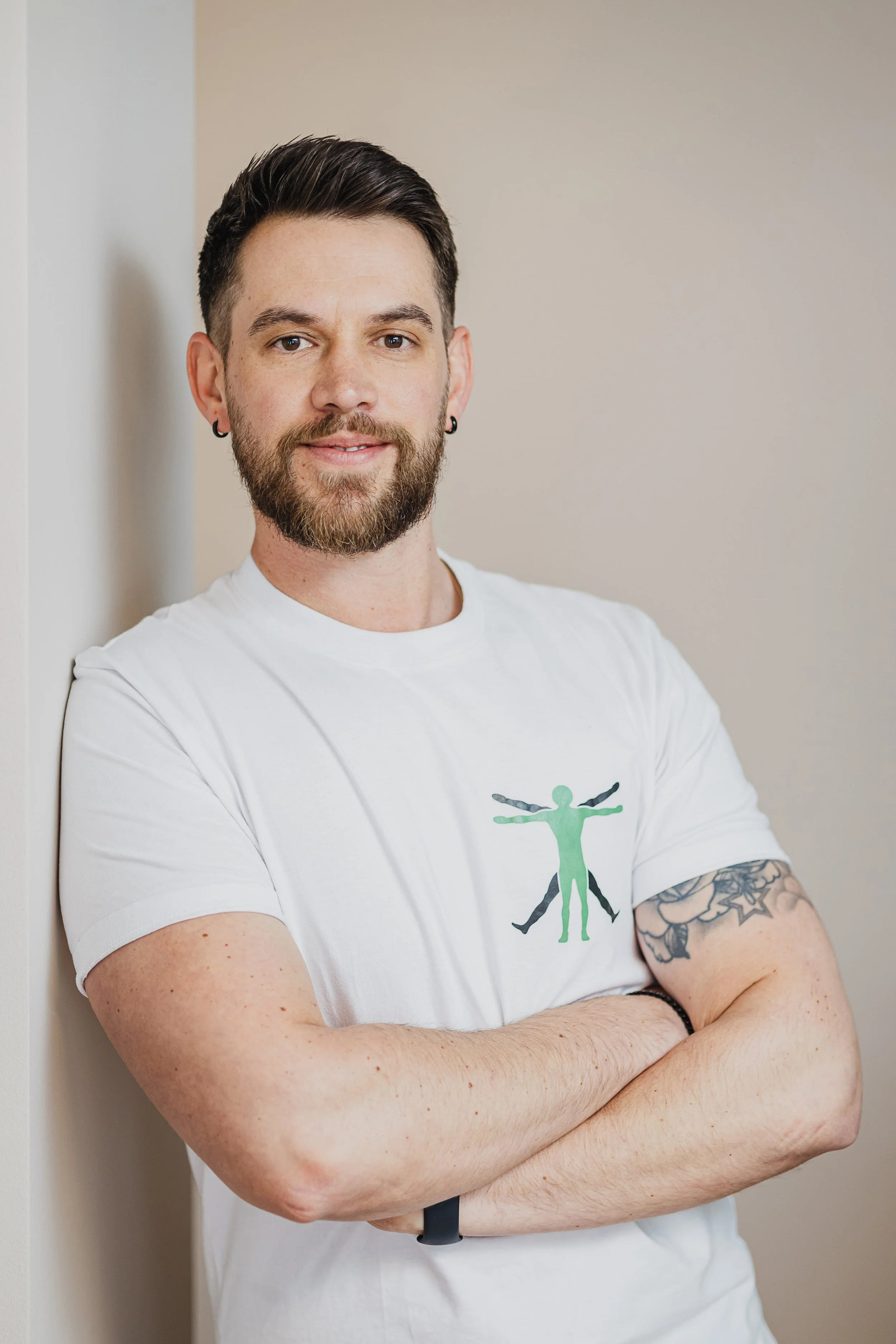 Junger Mann mit dunklem Haar, Bart, Ohrringen, in einem weißen T-Shirt, das ein menschliches Figur-Design zeigt, steht mit verschränkten Armen vor weißer Wand.