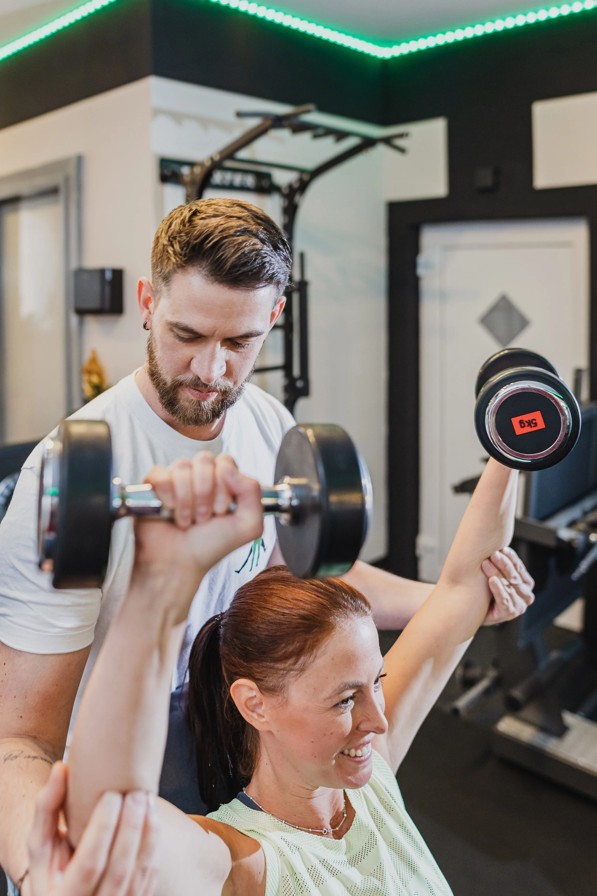 Zwei Frauen und ein Mann beim Gewichtheben im Fitnessstudio, der Mann hebt eine Hantel, die Frauen unterstützen. Im Hintergrund schwarzer Trainingsbereich mit Turngeräten und grüner LED-Beleuchtung an der Decke.