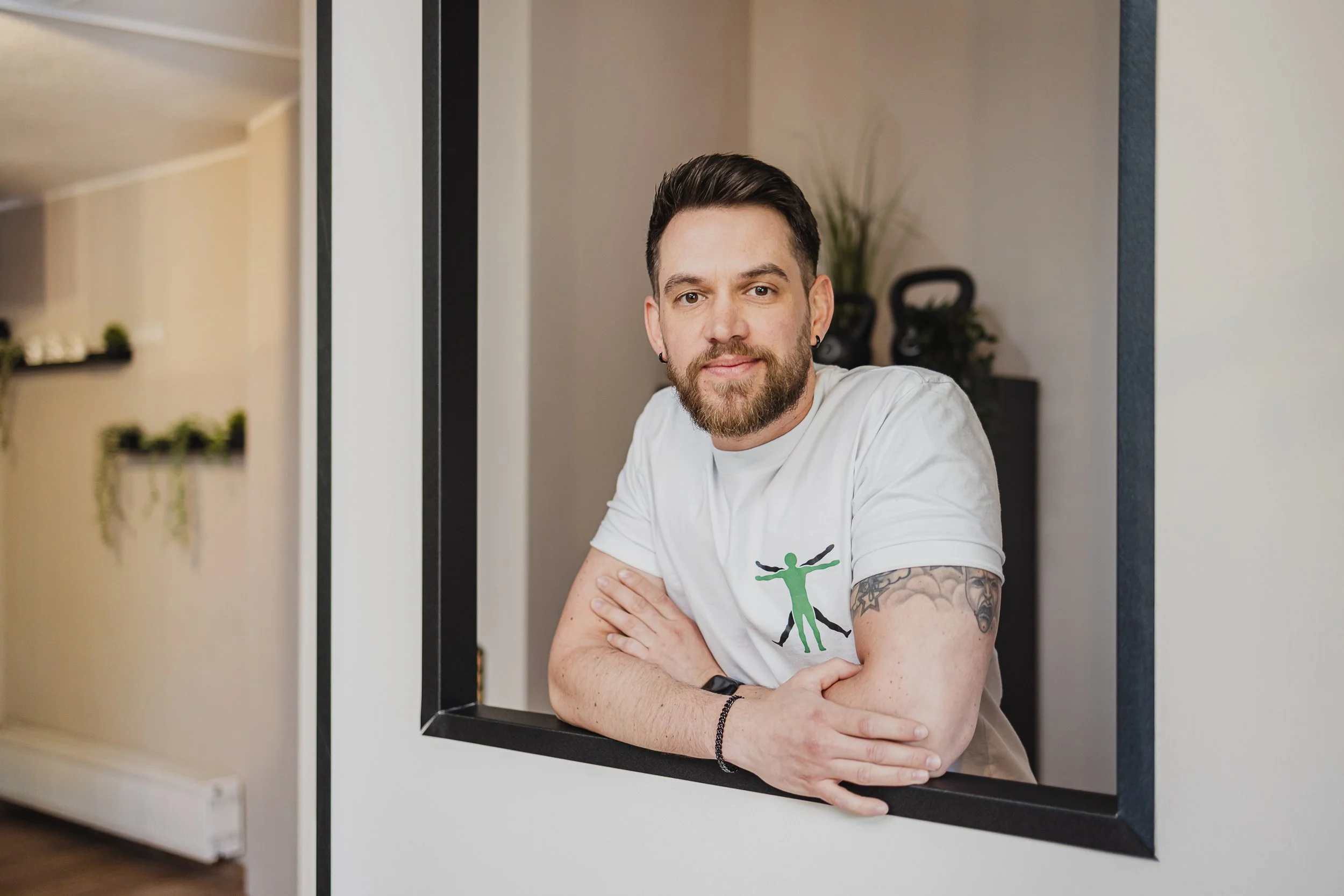 Ein Mann mit dunklen Haaren und Bart, trägt ein weißes T-Shirt mit einem grünen Mensch-Illustration, sitzt an einem Tisch, erscheint durch eine quadratische Öffnung in der Wand, im Hintergrund Pflanzen und Gymnastikgewichte.