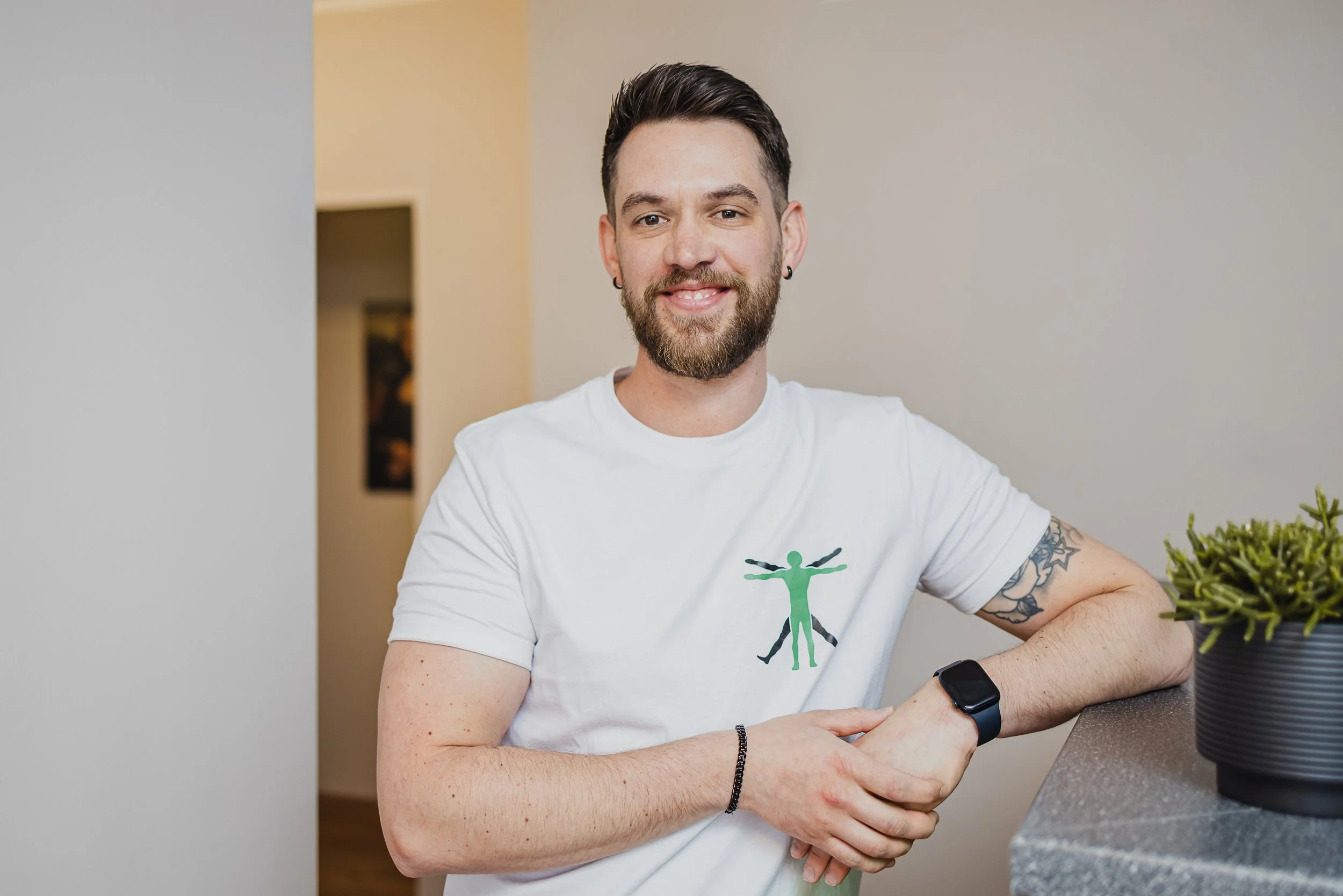 Junger Mann mit dunklem Haar, Bart und Tattoos, trägt weißes T-Shirt mit grüner Figur, sitzt an einem Tisch und lächelt, während er eine smartwatch trägt, im Hintergrund eine helle Wand.