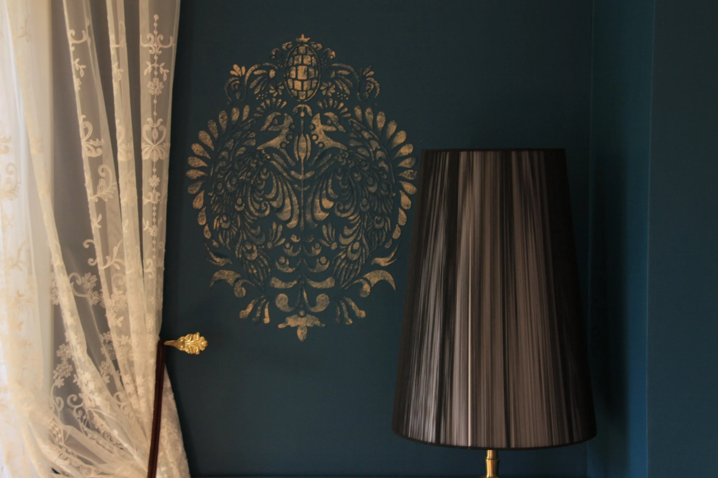 Blaue Wand mit goldener, ornamentaler Wanddekoration, vor einem schwarzen Lampenschirm, neben weißen, bedruckten Gardinen.