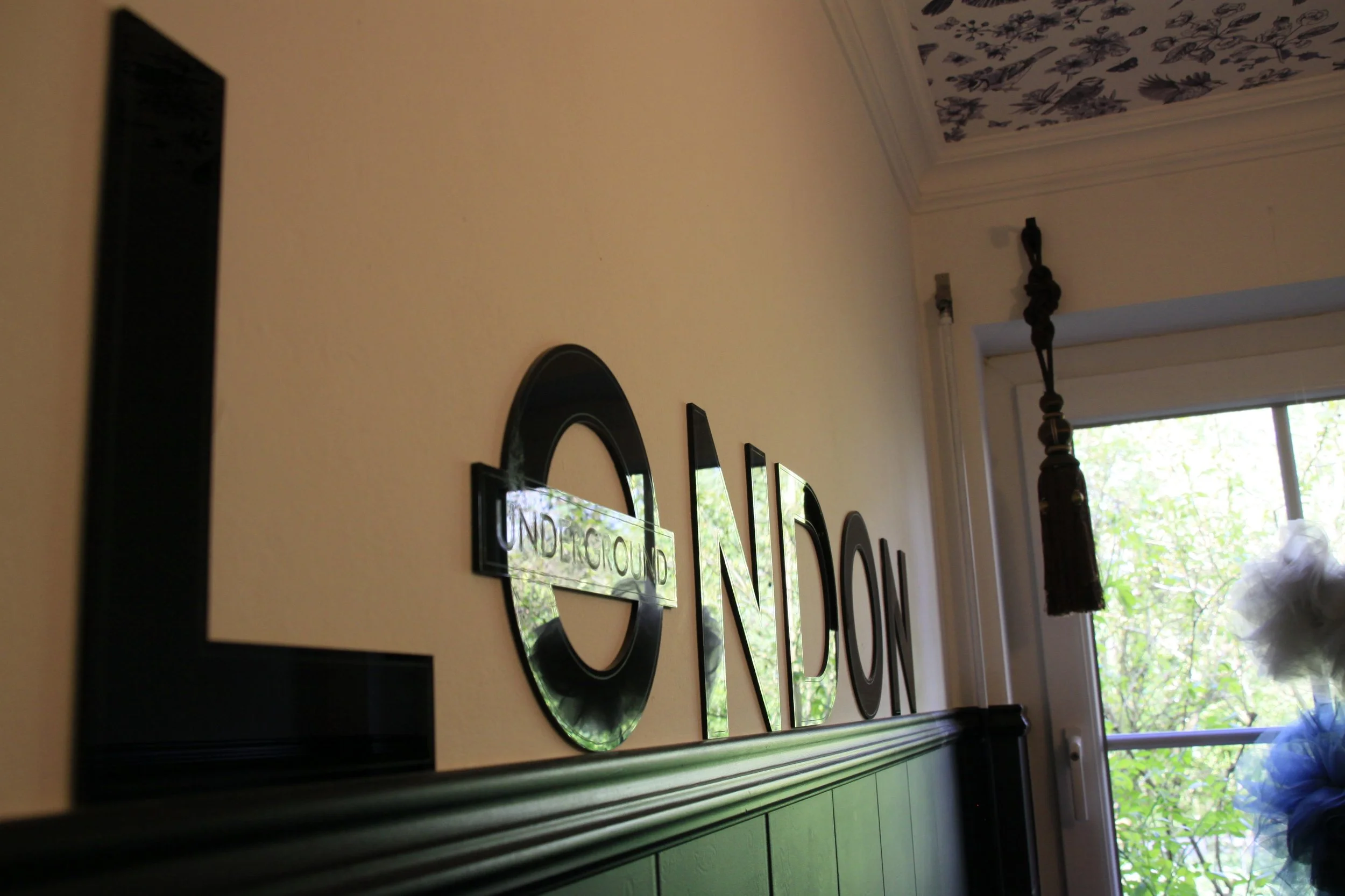 Der Blick auf eine Wand mit Spiegeln und einem Wandbild mit der Aufschrift 'UNDERGROUND' in einem Raum mit einer großen Fensteröffnung, durch die Bäume draußen sichtbar sind.