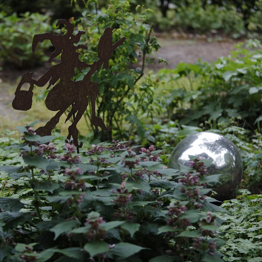 Ein Garten mit Pflanzen, einer metallenen dekorativen Skulptur eines Mannes mit einer Gießkanne und einem großen glänzenden silbernen Ball.