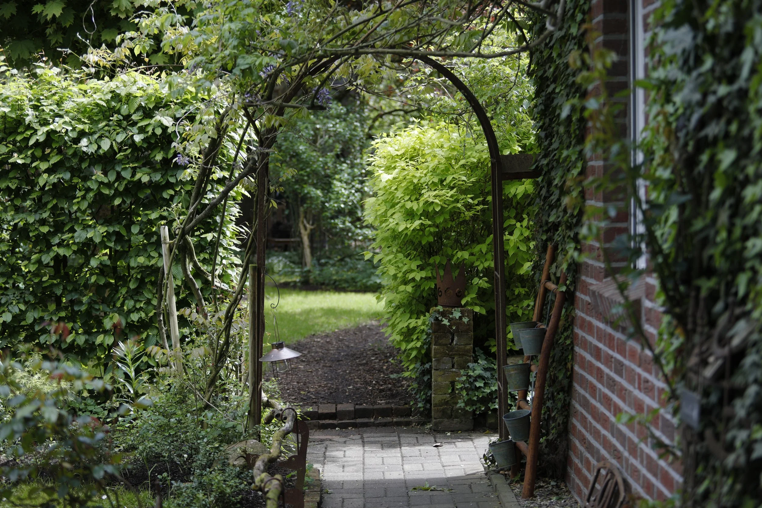 Ein Gartenweg, umgeben von grünen Pflanzen und einem roten Backstein-Haus. Ein Torbogen aus Metall mit Pflanzen darüber bildet den Eingang zu einem Garten. Es ist tagsüber, das Licht ist sanft.