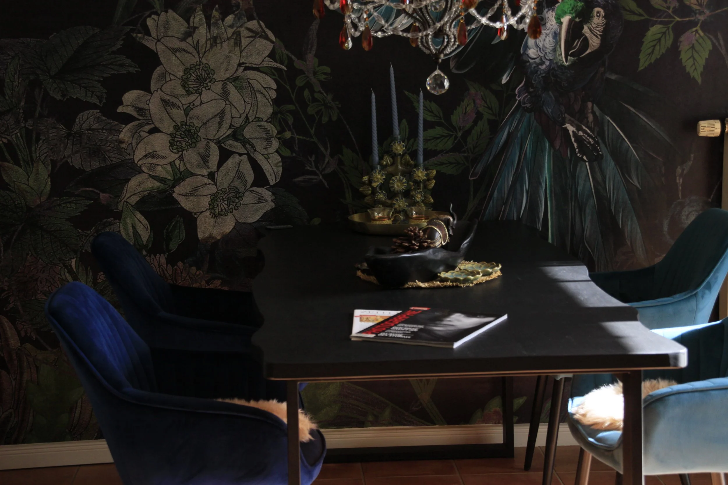 Ein dunkler Raum mit einer schwarzen Wand, die mit großen weißen Blüten und grünen Blättern bedruckt ist. Es gibt einen runden Tisch mit Sektempfangs- und Blumenarrangements. Mehrere blaue und graue Stühle sind um den Tisch gruppiert. An der Decke hängt eine Kristalllampe.