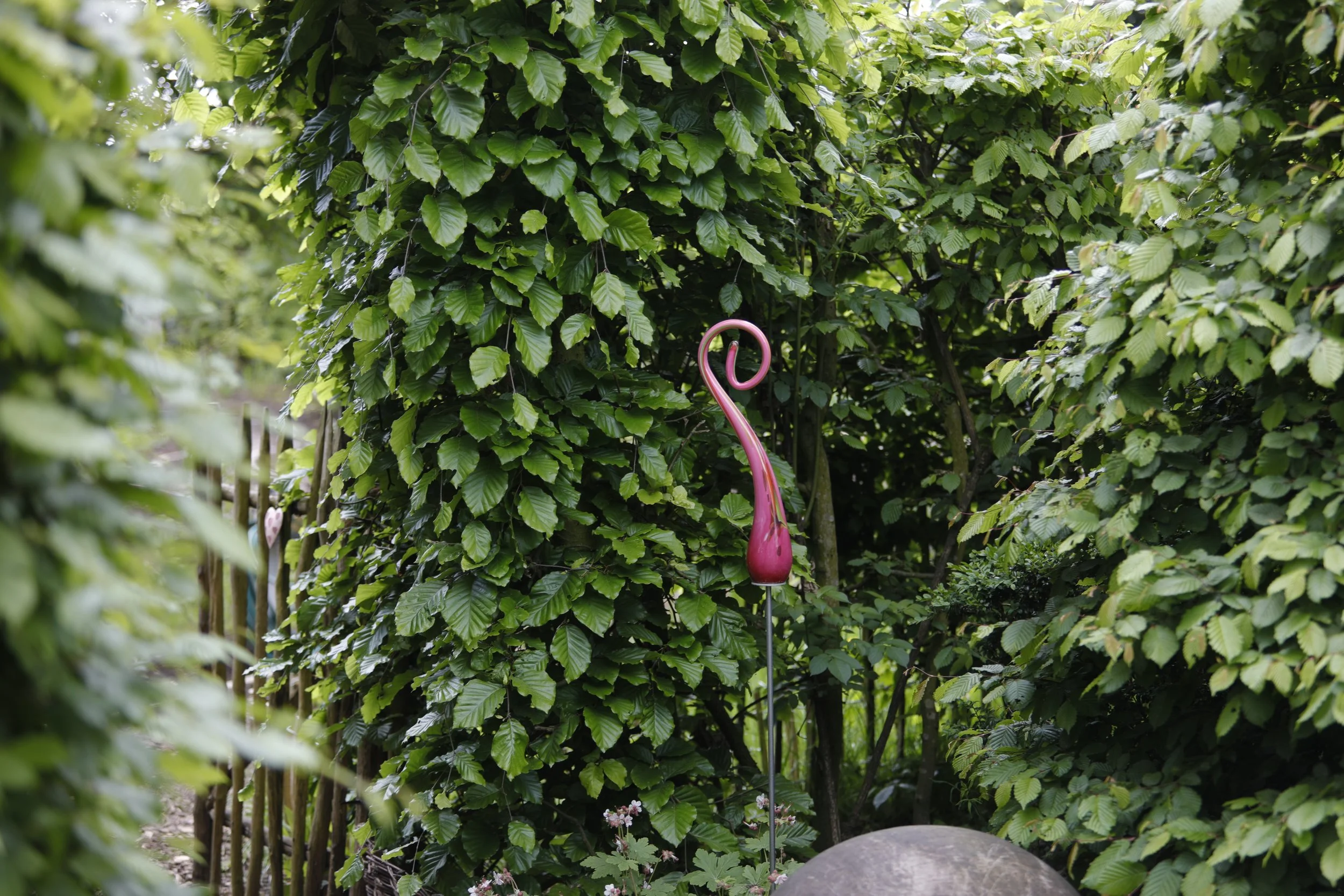 Ein Garten mit grünen Blättern, in dem ein pinkfarbener Flamingo-Dekorationsstab steht. Im Vordergrund ist ein großer Felsen zu sehen, und im Hintergrund sind einige kleine Blumen sichtbar.