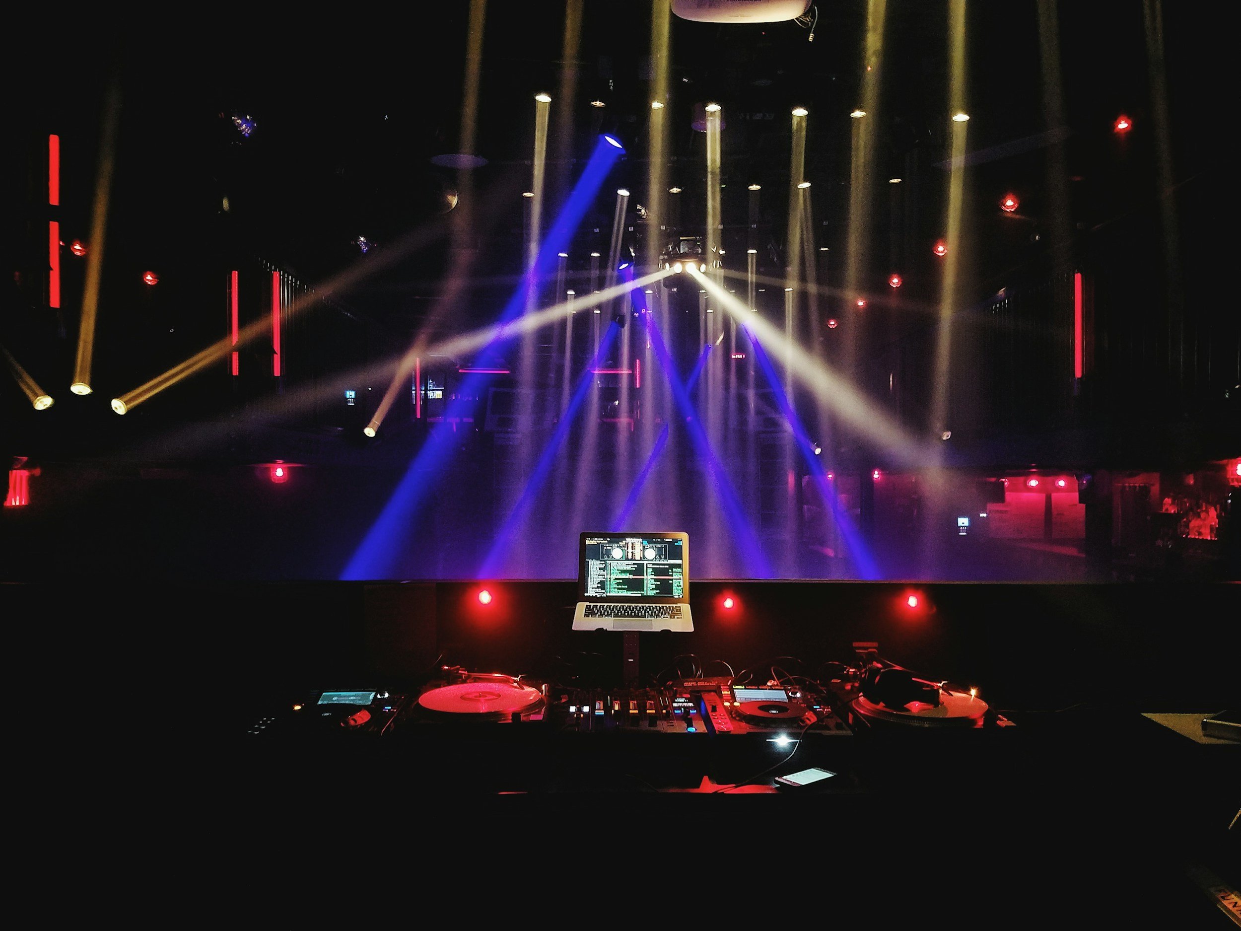 DJ equipment on a nightclub stage with colorful lights and fog, empty dance floor in the background.