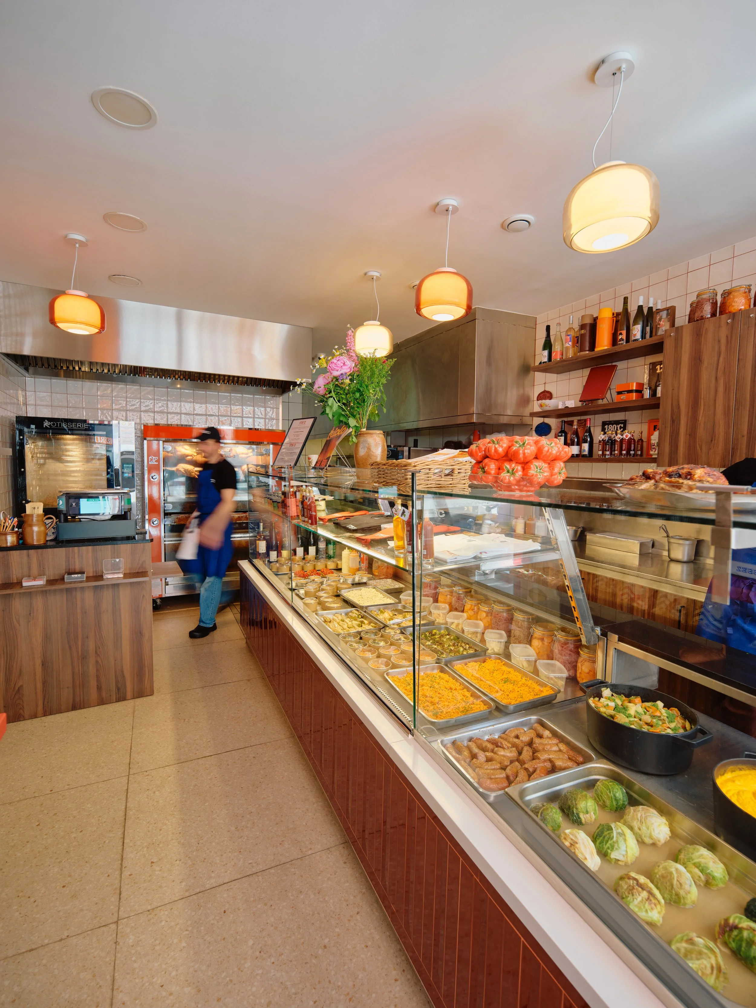 Vitrine de nourriture dans un restaurant, avec des plats variés comme des légumes, des pommes de terre, des sauces, et une décoration avec un bouquet de fleurs et des bouteilles sur une étagère en bois.