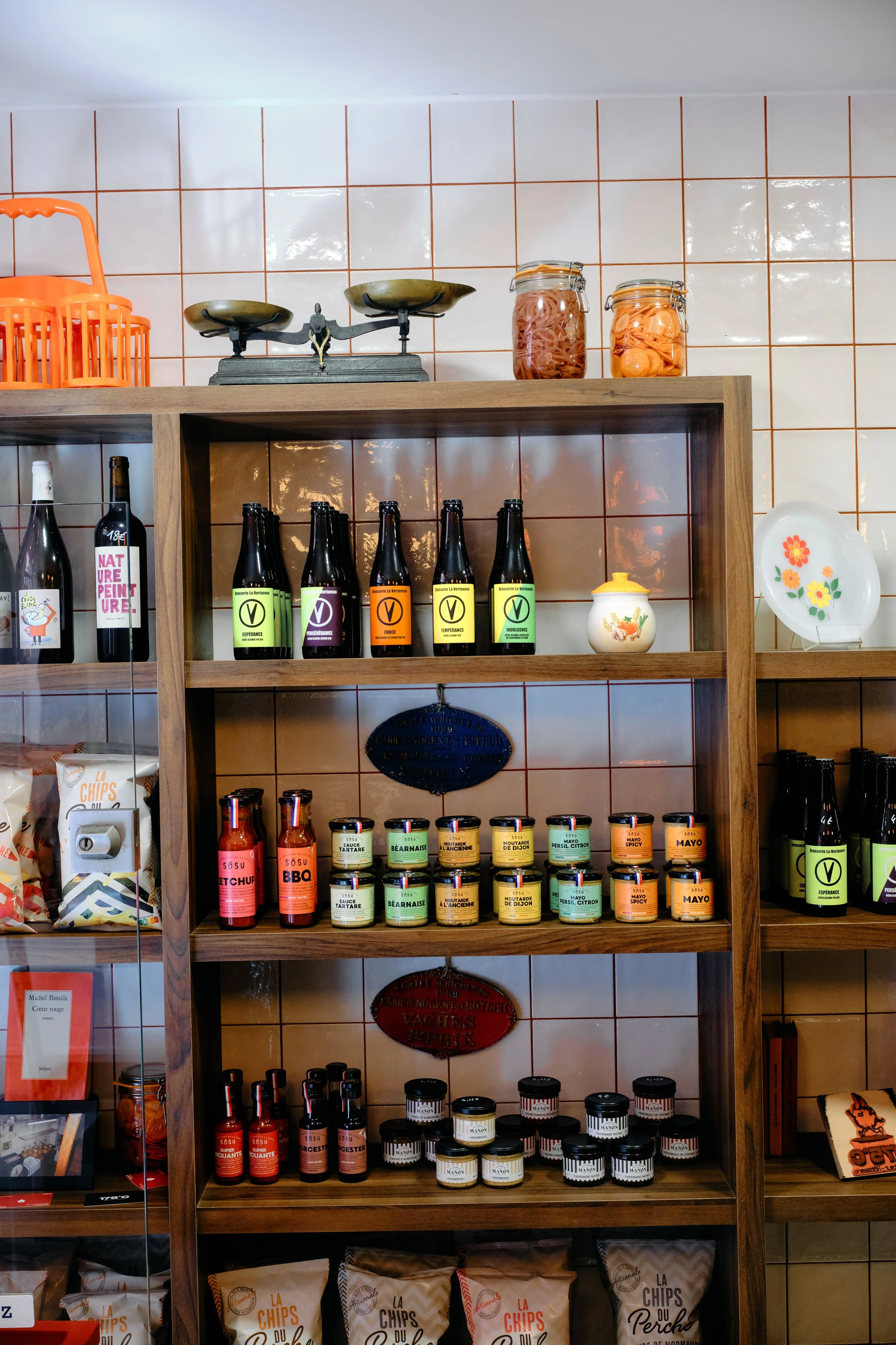 Étagère en bois avec diverses bouteilles, bocaux d'épices, conserves, et objets de décoration sur un mur de carreaux blancs avec des lignes orange.