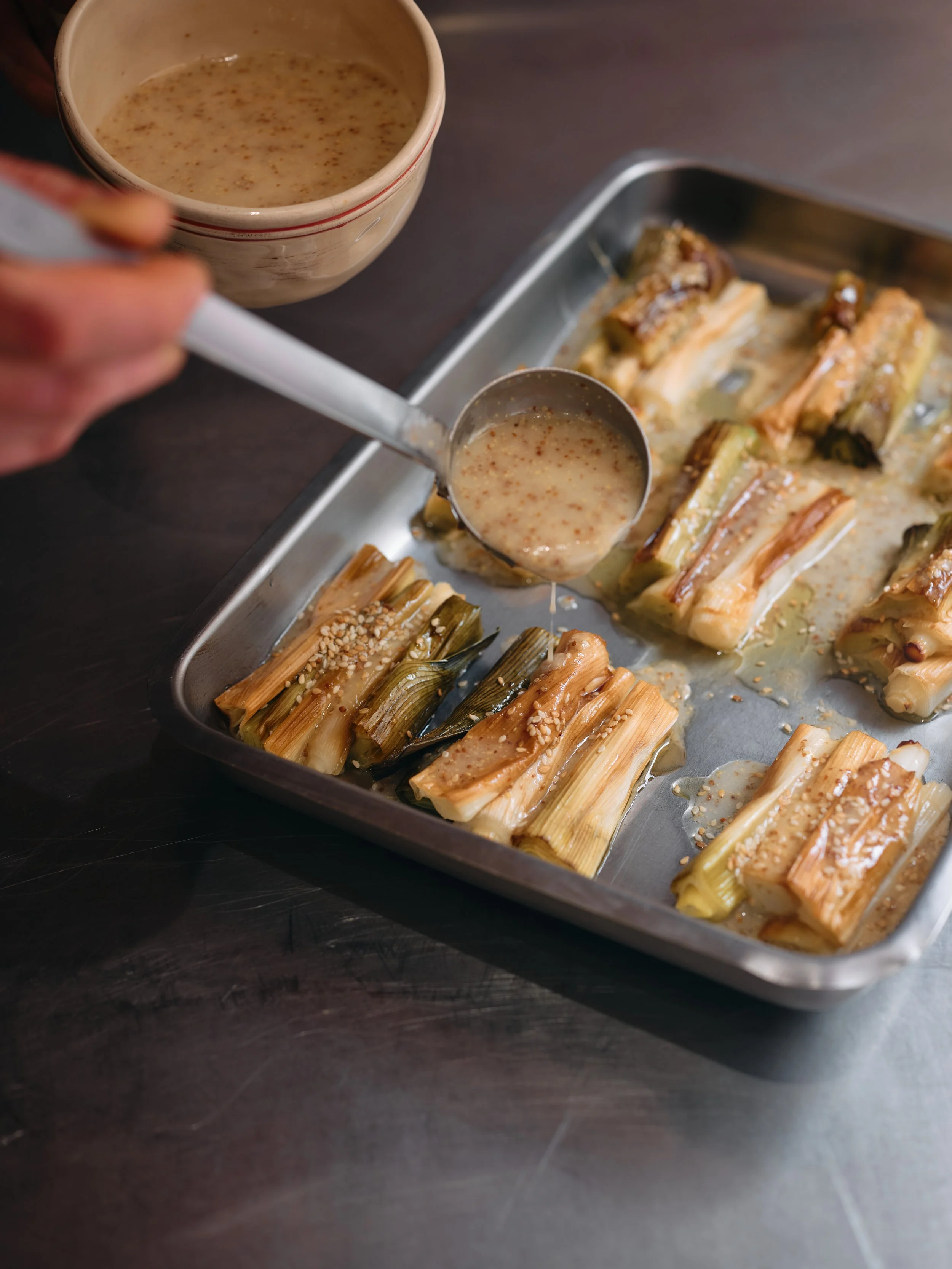 Des rouleaux de poireaux, dans un plat en métal, avec une cuillère contenant une sauce vinaigrette maison.