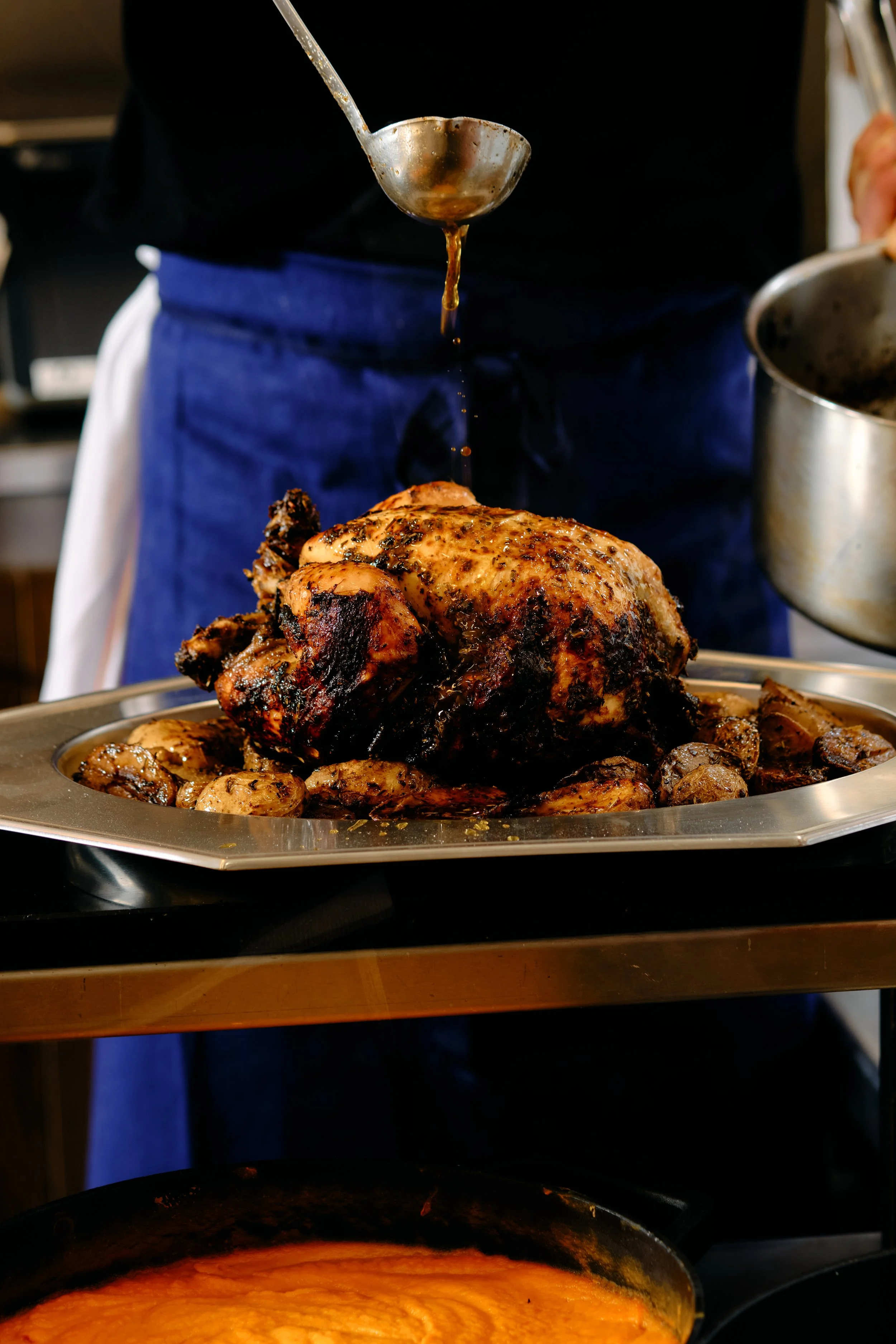 Une plaque de cuisson avec un poulet rôti entouré de pommes de terre, deux casseroles en arrière-plan, et une sauce ou un jus coulant d'une cuillère suspendue.