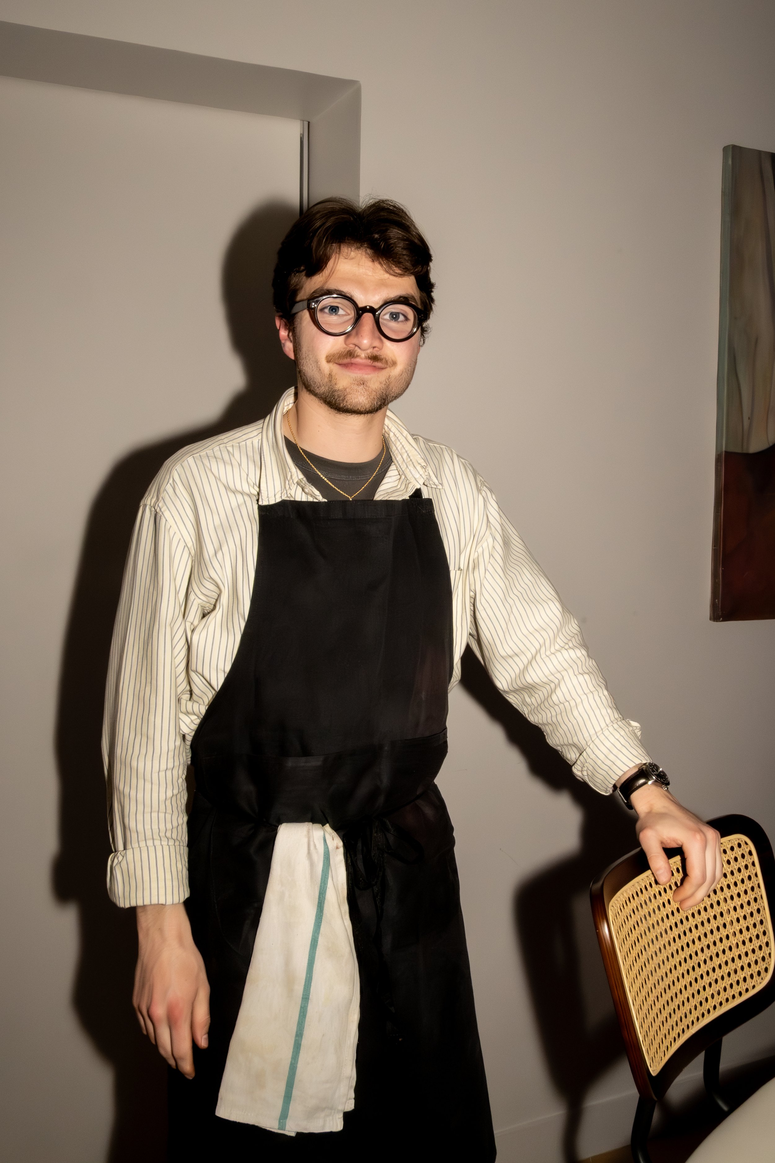 Un homme avec des lunettes, une barbe, portant une chemise à rayures, un tablier noir, et un bracelet, se tenant près d'une chaise en bois, dans une pièce avec un mur blanc.