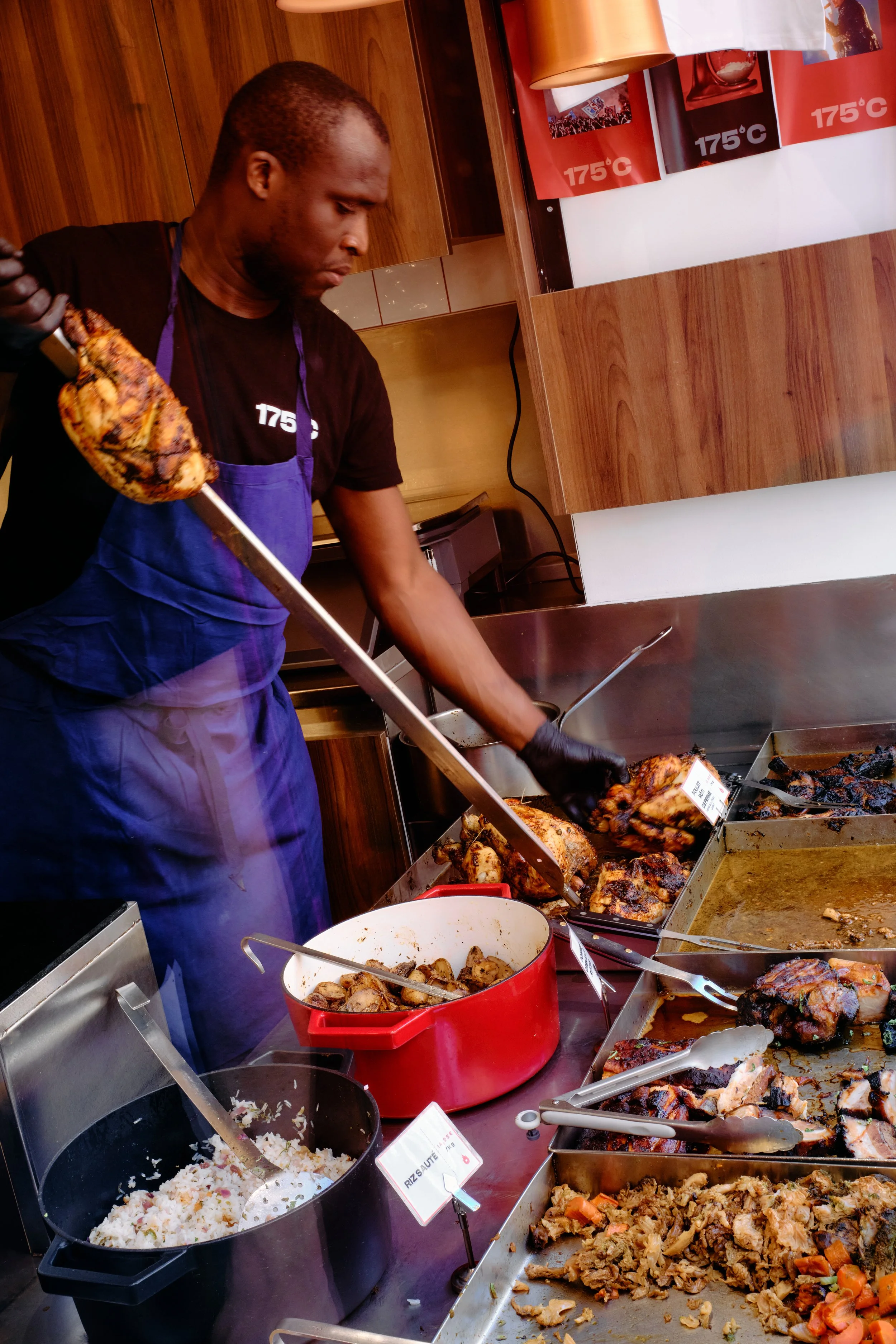 Un homme prépare de la nourriture dans une cuisine, avec divers plats et aliments, comme du riz, du poulet rôti, et un mélange de légumes, sur un comptoir en acier.