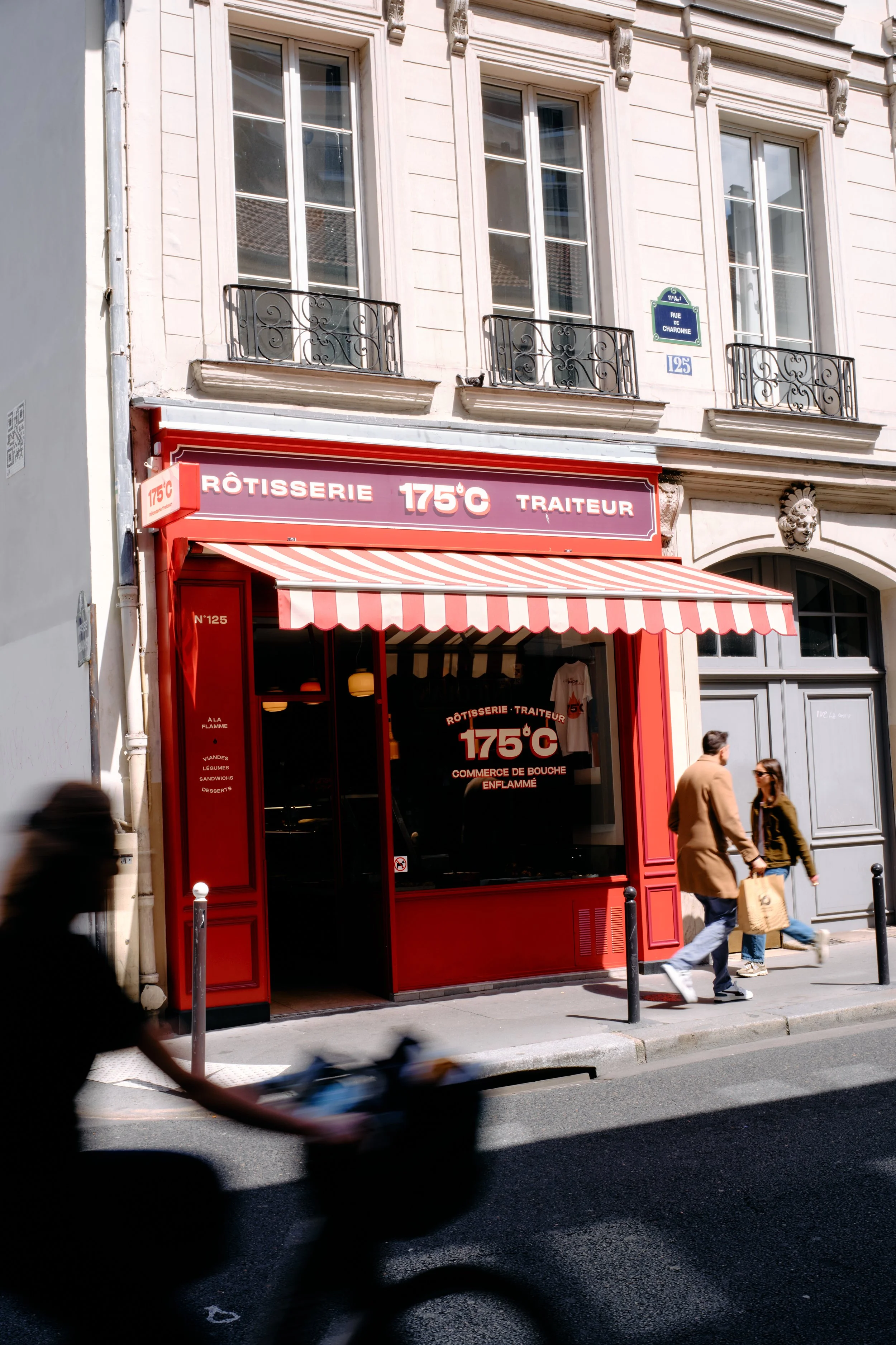 Façade d'une rôtisserie et traiteur, avec une enseigne mentionnant 175°C, située au 125 rue de Charonne dans un immeuble haussmannien avec trois fenêtres à balustres en fer forgé au premier étage. Deux personnes, un homme et une femme, marchent devant la boutique, en passant devant la vitrine et une personne passe rapidement devant la caméra à gauche, floue.
