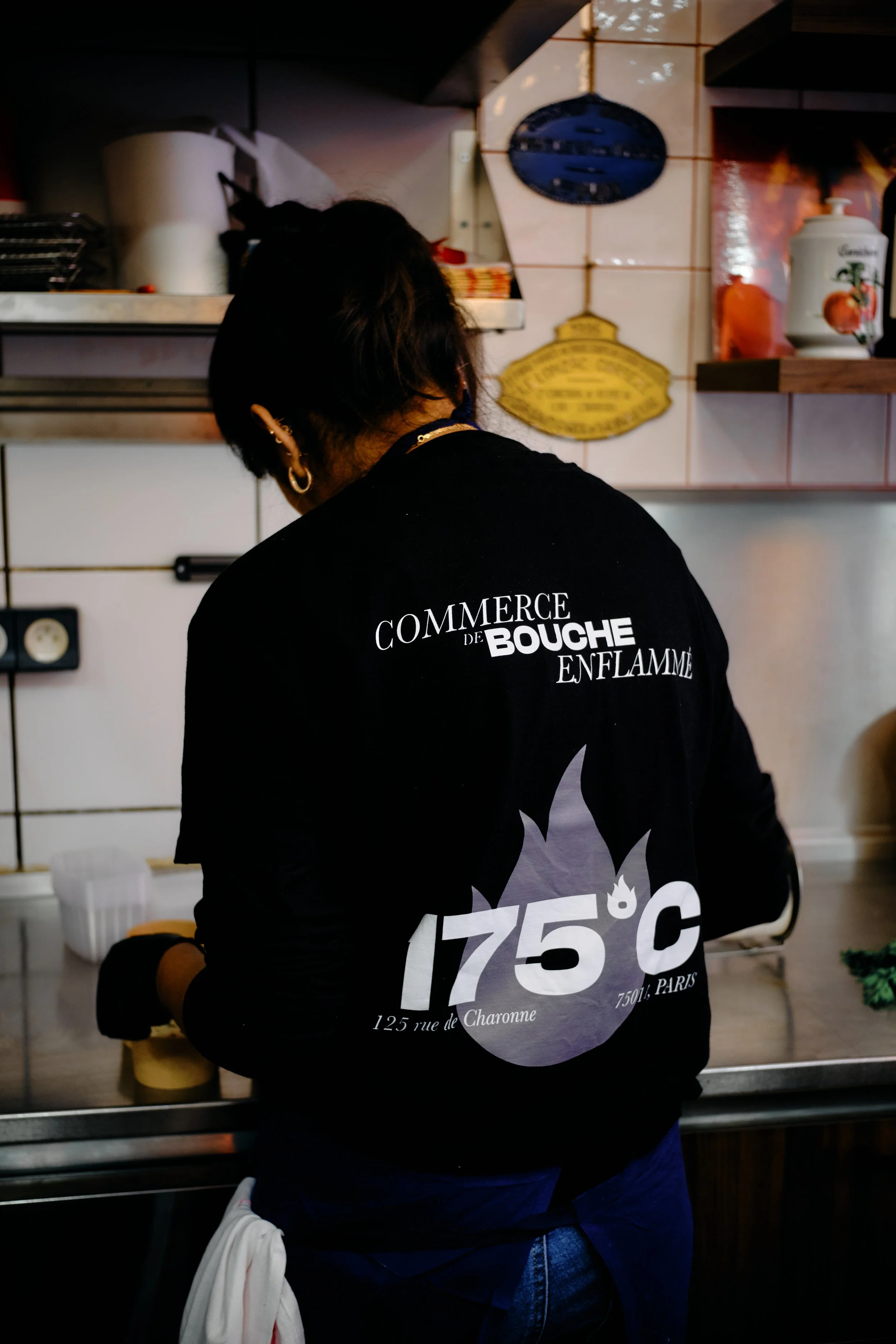 Personne portant un sweat noir avec écritures en français, cuisinière en arrière-plan, cuisine avec des tuiles blanches, étagère en bois et objets de cuisine.
