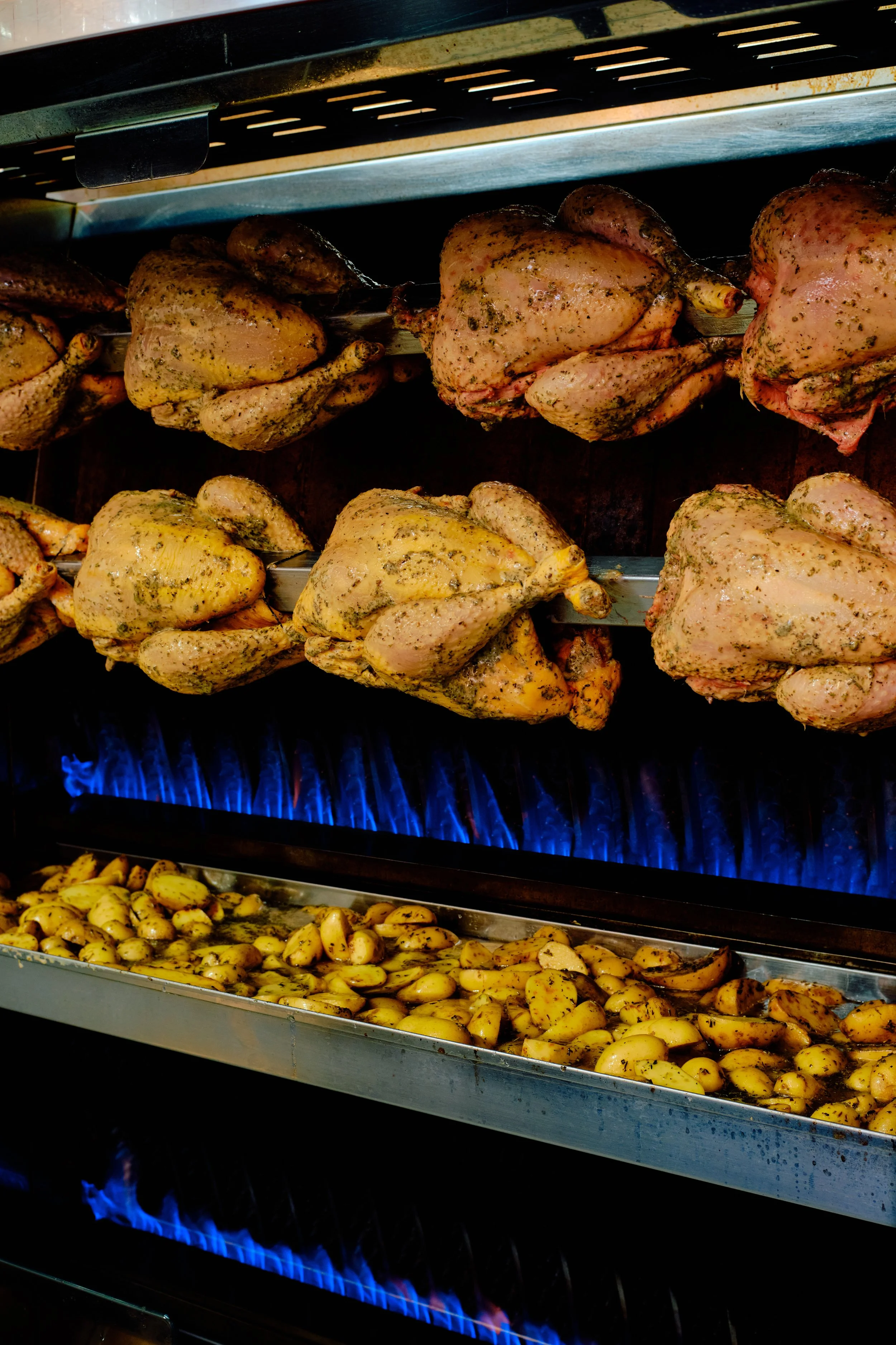 Poulet rôti épicé en train de cuire sur une broche dans un four, avec des pommes de terre assaisonnées dans une grande plaque en dessous, sous une lumière de cuisson bleue.