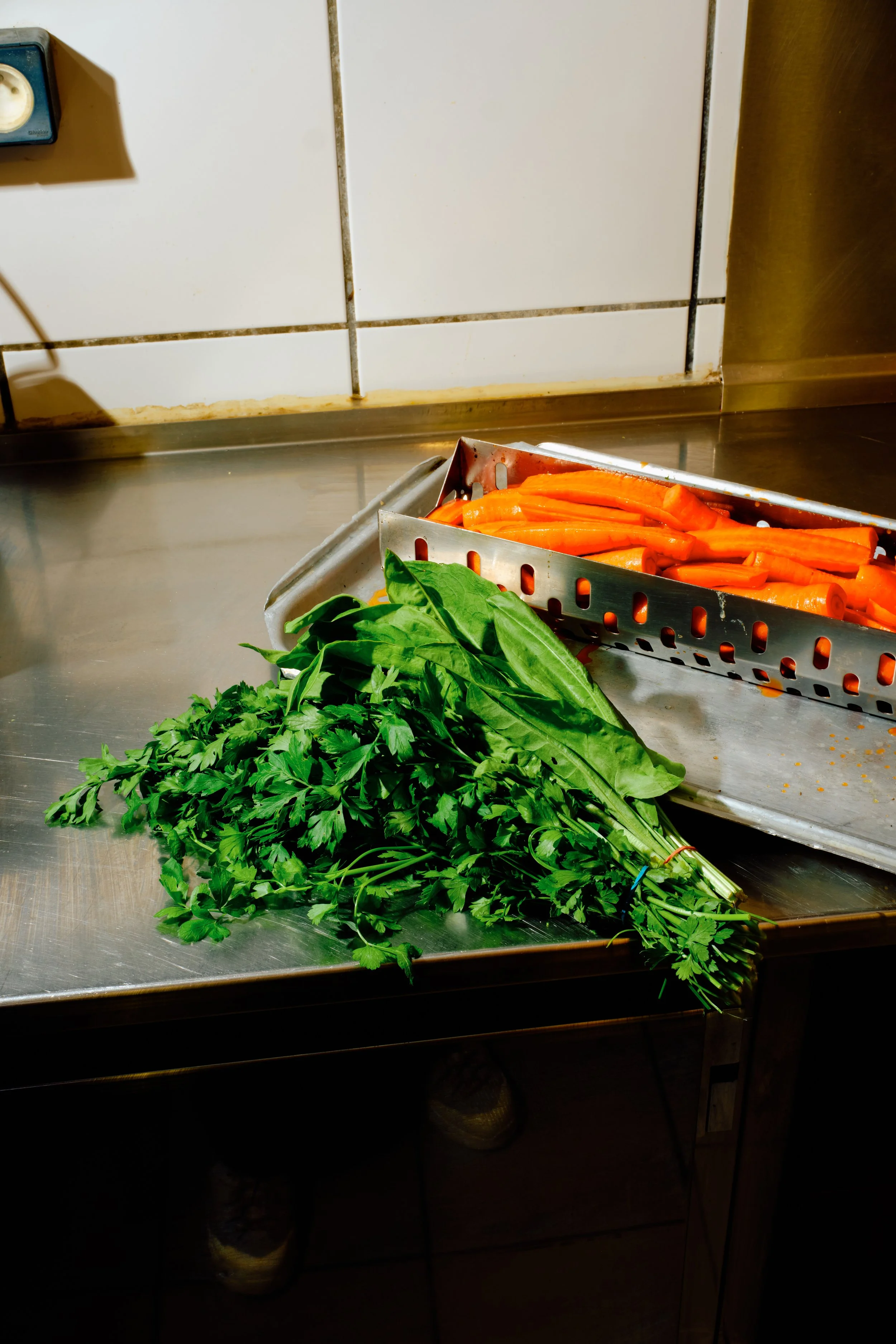 Des carottes, du basilic, du persil et de la ciboulette posés sur un plan de travail en cuisine, avec un panier en métal contenant des carottes coupées en tronçons.