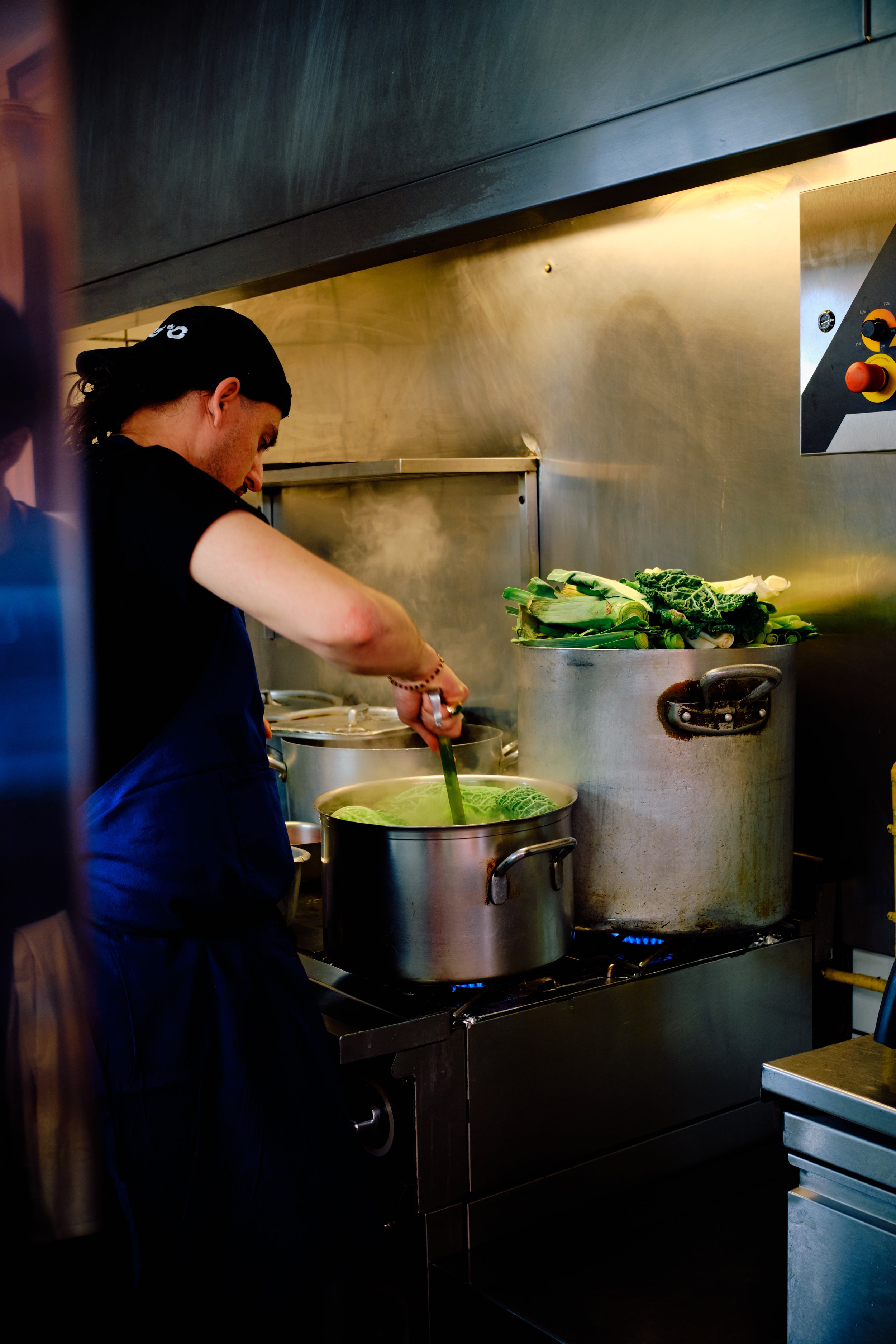 Une personne en cuisine, préparant des légumes verts dans une grande casserole sur une cuisinière industrielle, à l'arrière-plan un mur en acier inoxydable avec commandes