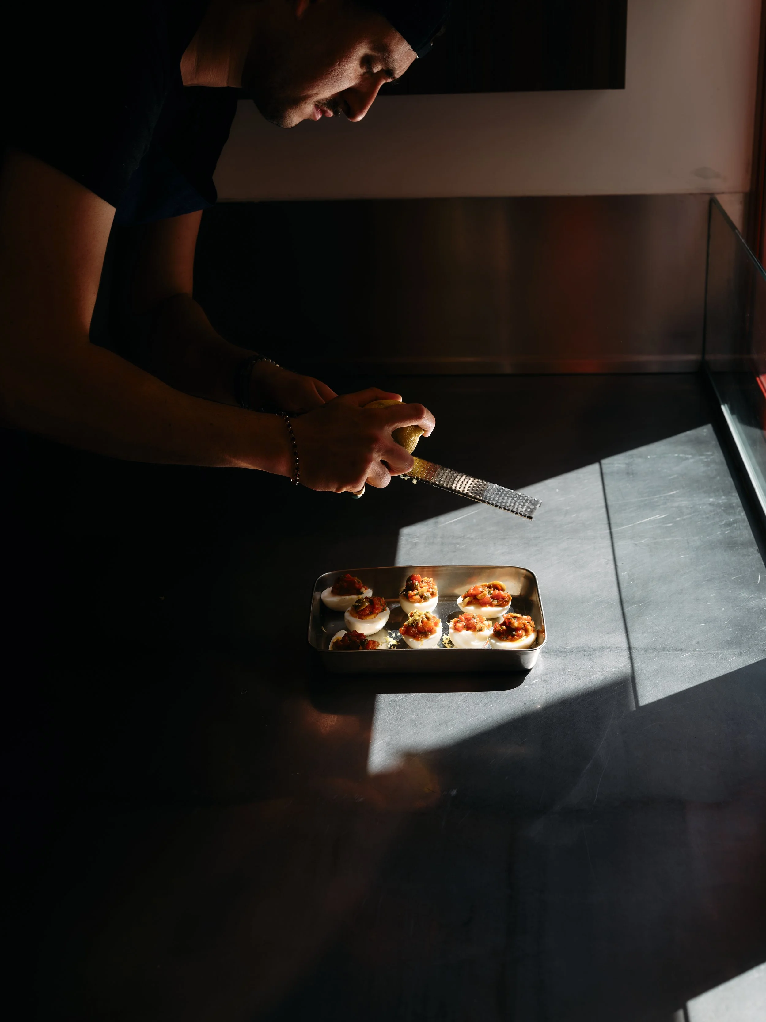 Un homme en noir prépare des œufs farcis avec une râpe à fromage, sur une surface noire, près d'une fenêtre avec de la lumière naturelle