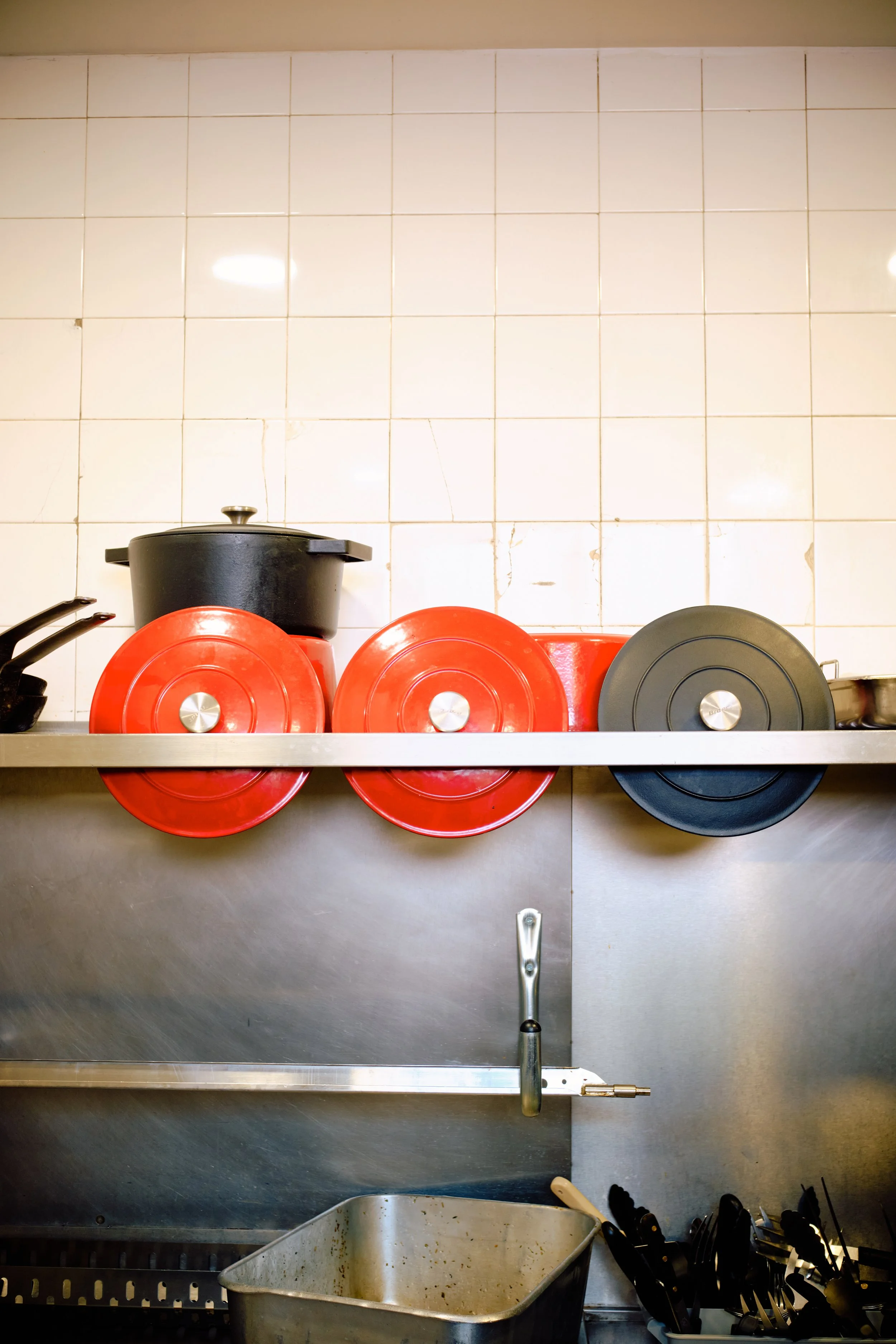 Une cuisine avec des casseroles rouges et noires, des couverts noirs, un évier en inox et un mur en carreaux blancs.