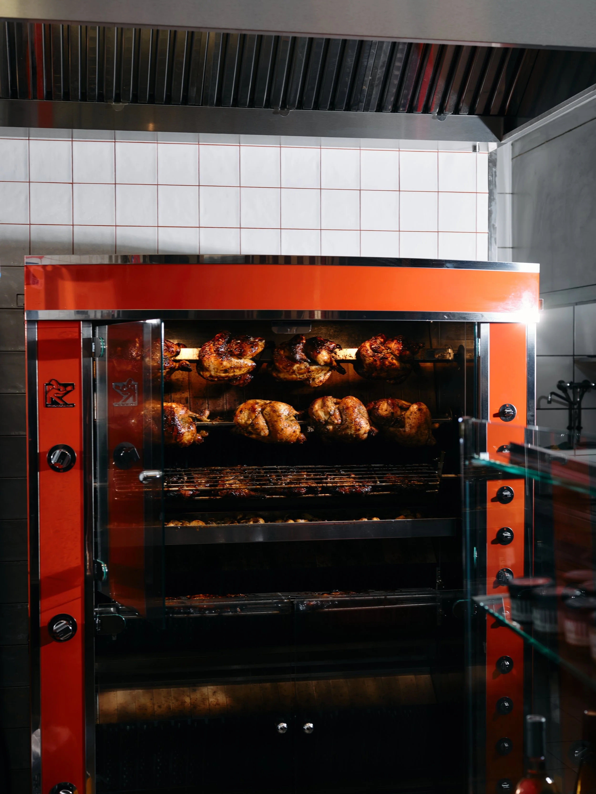 Poulet rôti cuit dans une rôtissoire à gaz, visible à travers une porte en verre d'une rôtissoire à gaz orange vif, avec un mur en carrelage blanc en arrière-plan.