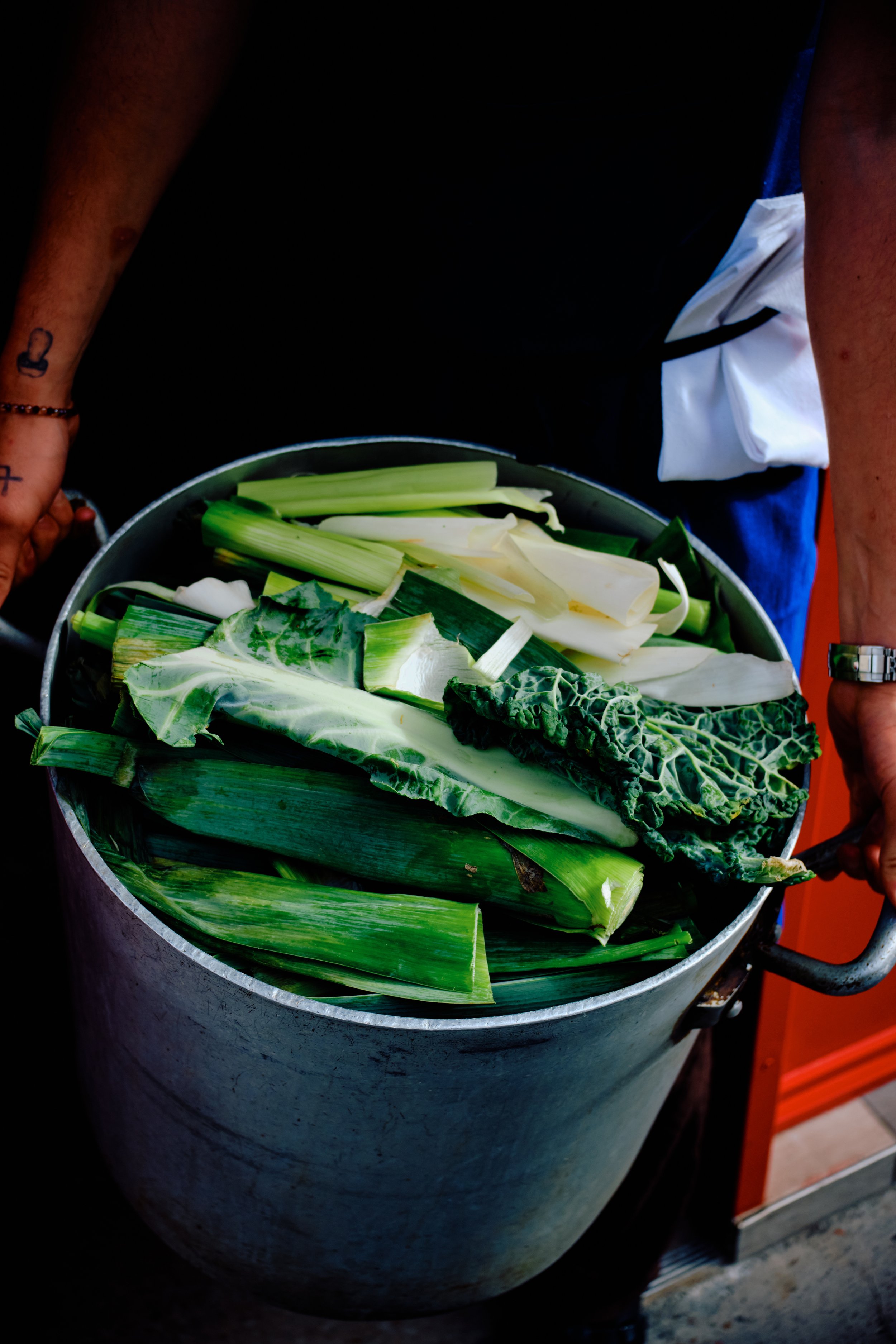 Une grande cuve contenant divers légumes verts, notamment du chou, de la ciboulette, et d'autres feuilles vertes, probablement pour la préparation culinaire.
