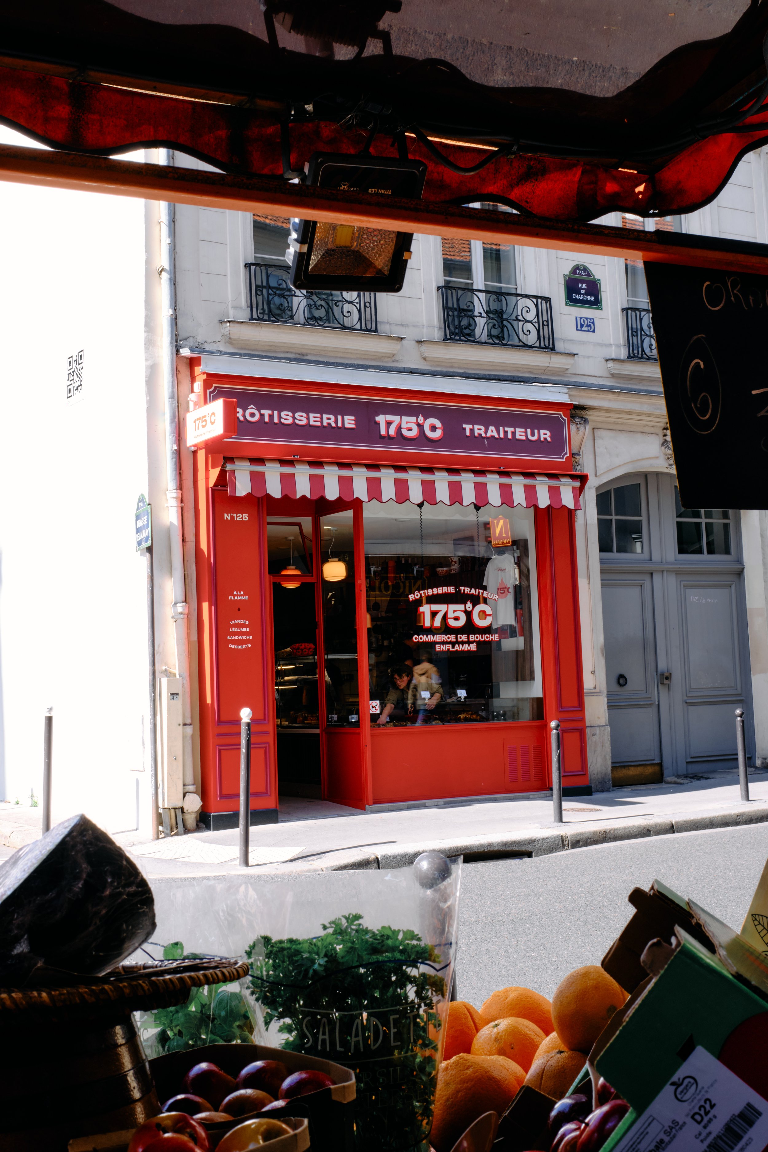 Vitrine d'un marché avec des fruits comme des oranges, des pommes et des cerises, vue depuis un stand à l'extérieur, avec une boulangerie appelée '175°C Traiteur' en arrière-plan.