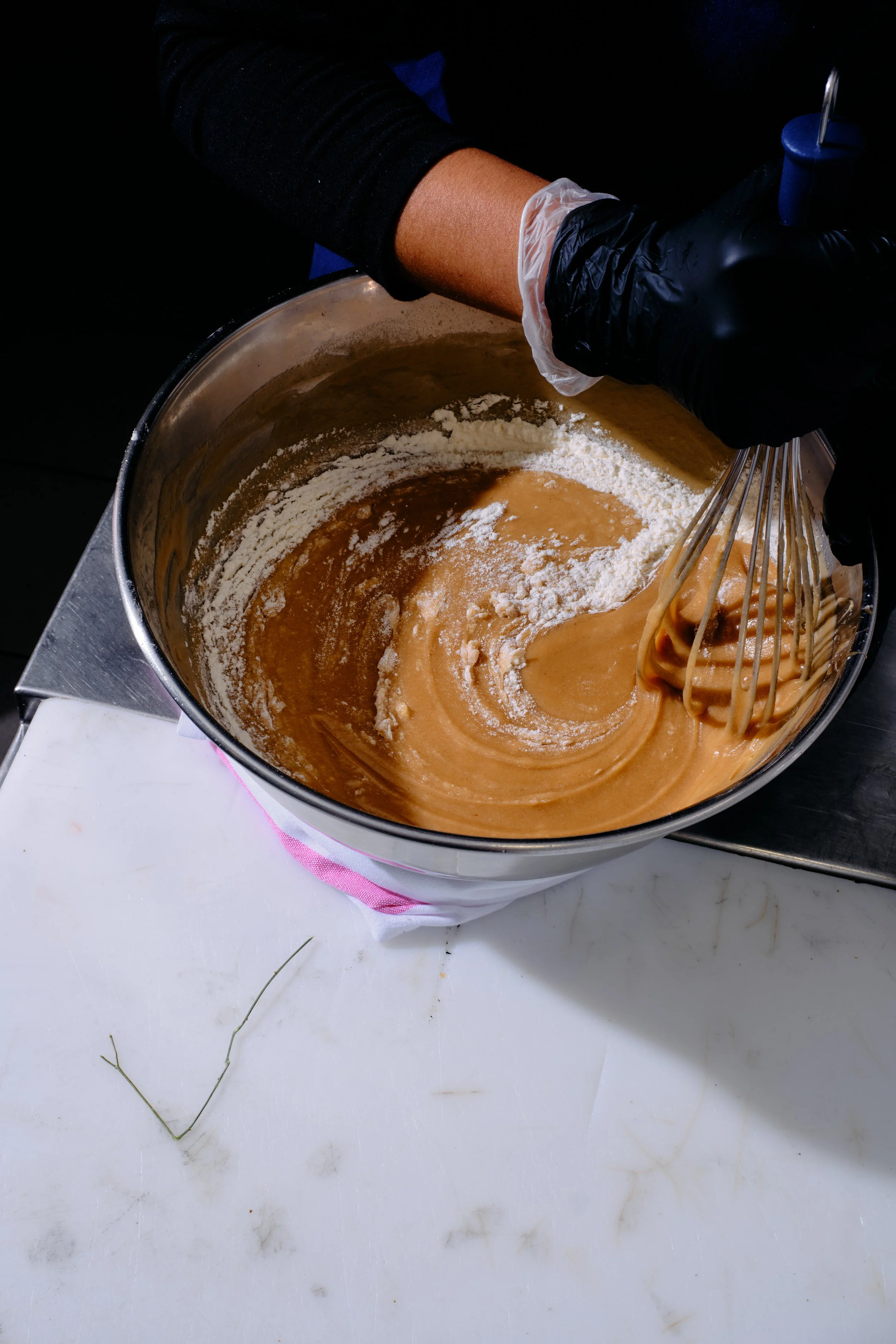 Mélangeur avec une main portant un gant noir mélangeant une pâte dans un bol en acier inoxydable, avec la main portant de la cireur en plastique blanc et un autre gant noir, sur une surface blanche.