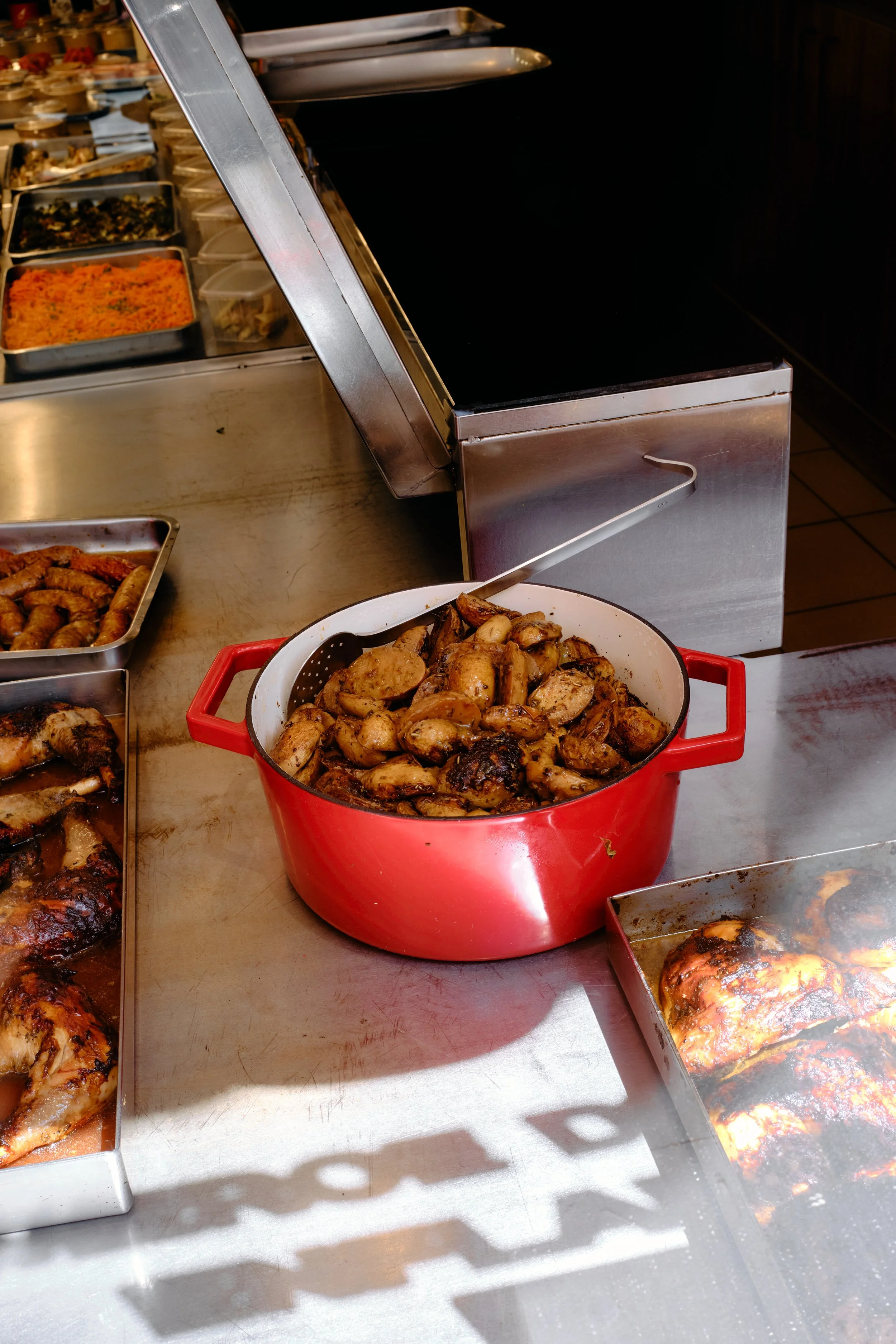 Plat en acier inoxydable avec diverses salades, une grande casserole rouge avec des champignons cuits, and une assiette avec du poulet grillé. Le fond montre une vitrine avec d'autres plats et aliments.