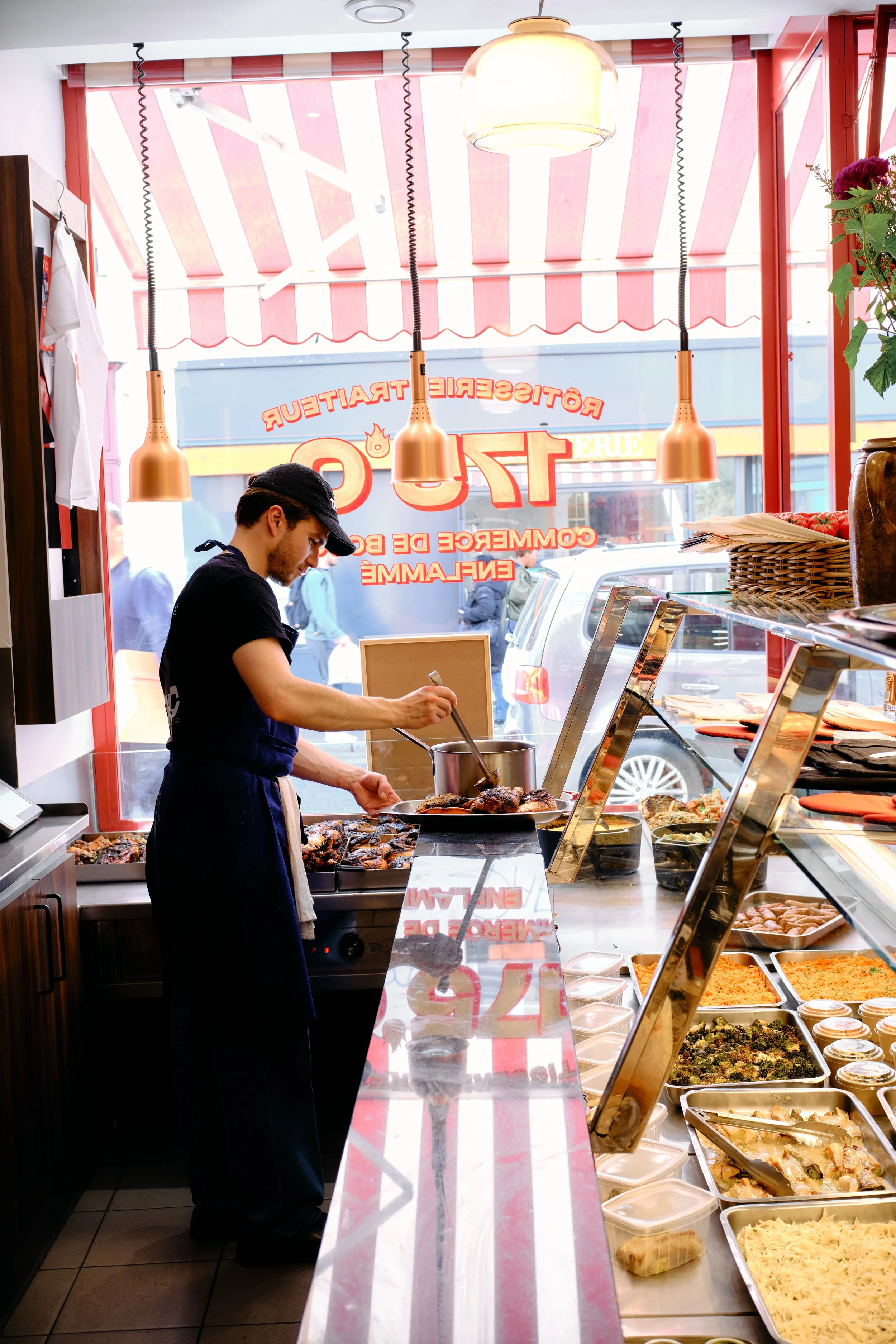 Un jeune homme prépare un plat devant une station de restauration avec une vitrine de plats chauds, incluant des salades et des accompagnements, à l'intérieur d'une boutique avec de grandes fenêtres et des enseignes extérieures visibles.