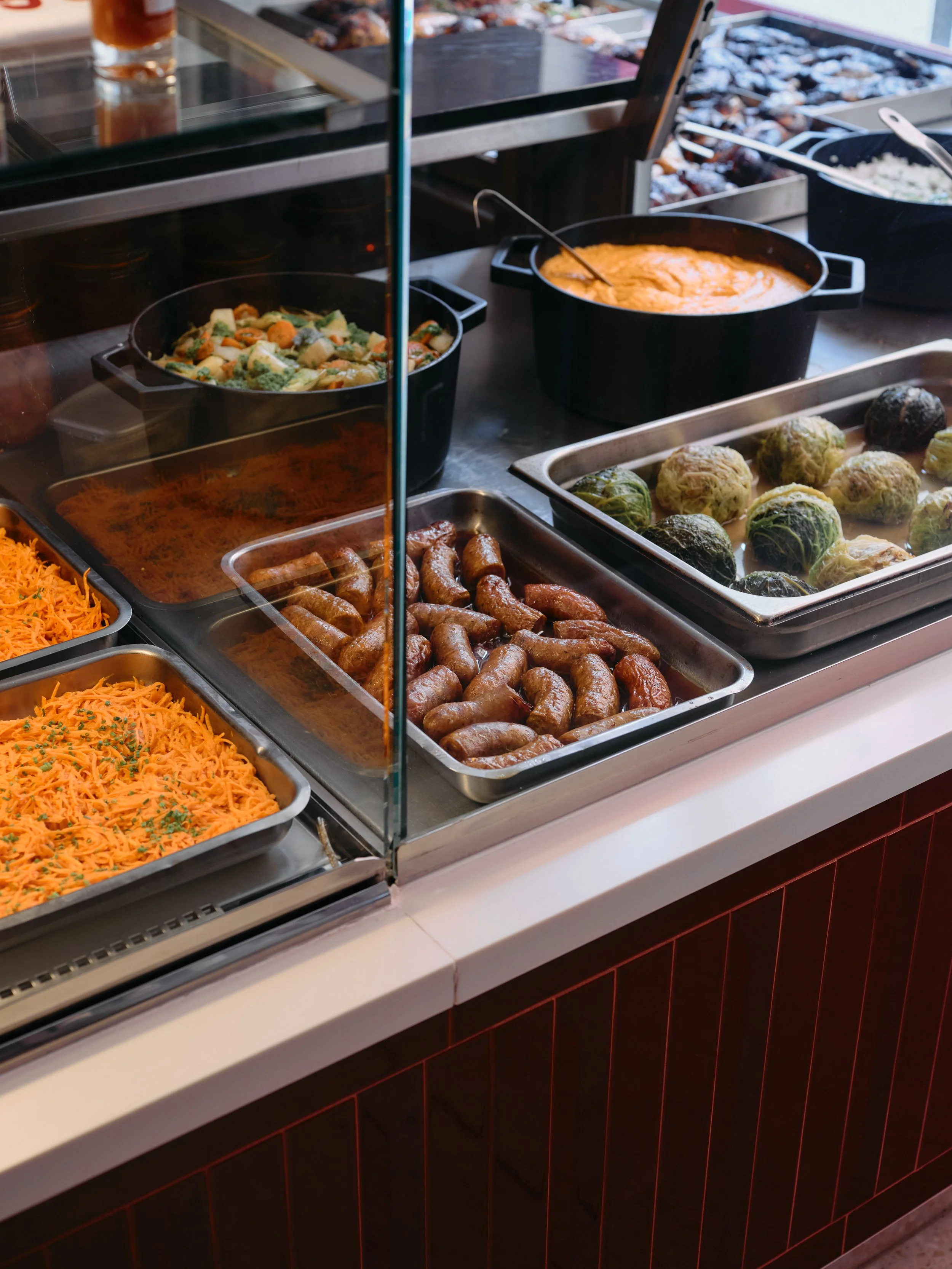 Buffet de plats chauds et froids, comprenant saucisses, légumes, riz et autres plats dans de grands contenants en métal, derrière une vitre de présentation.