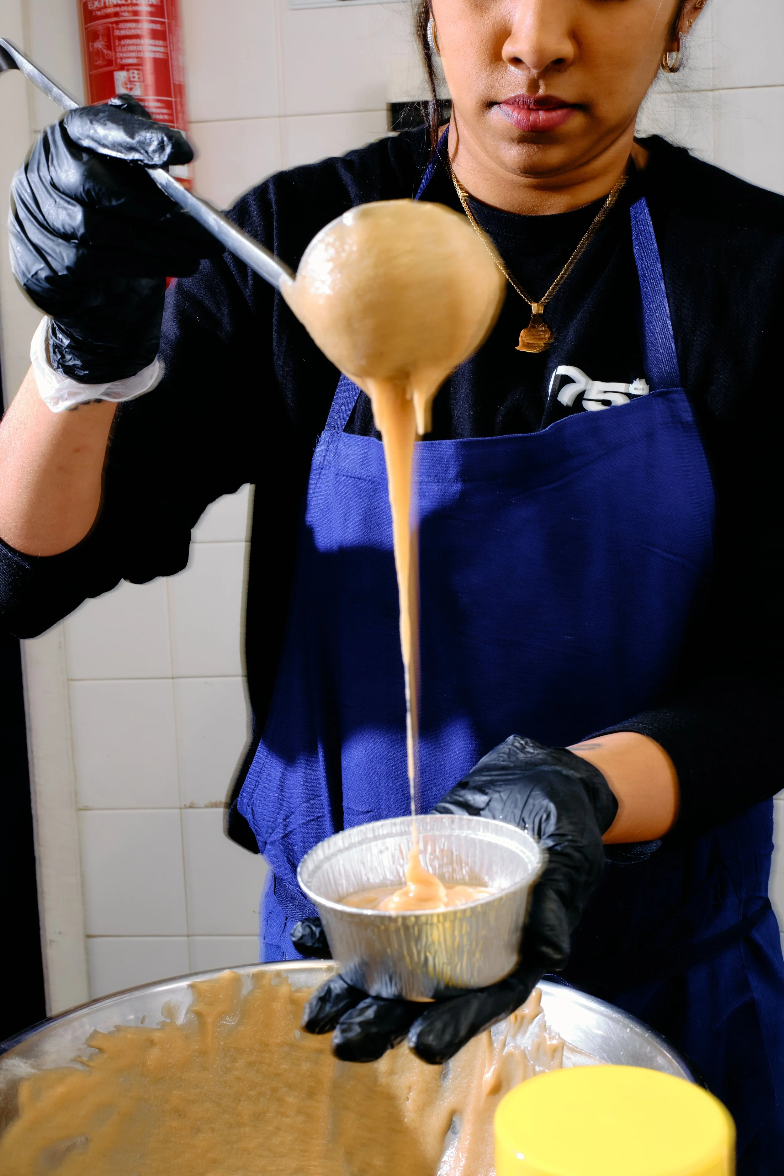 Une femme en noir, portant un bleu et un gant noir, verse de la pâte de caramel dans un récipient en aluminium.
