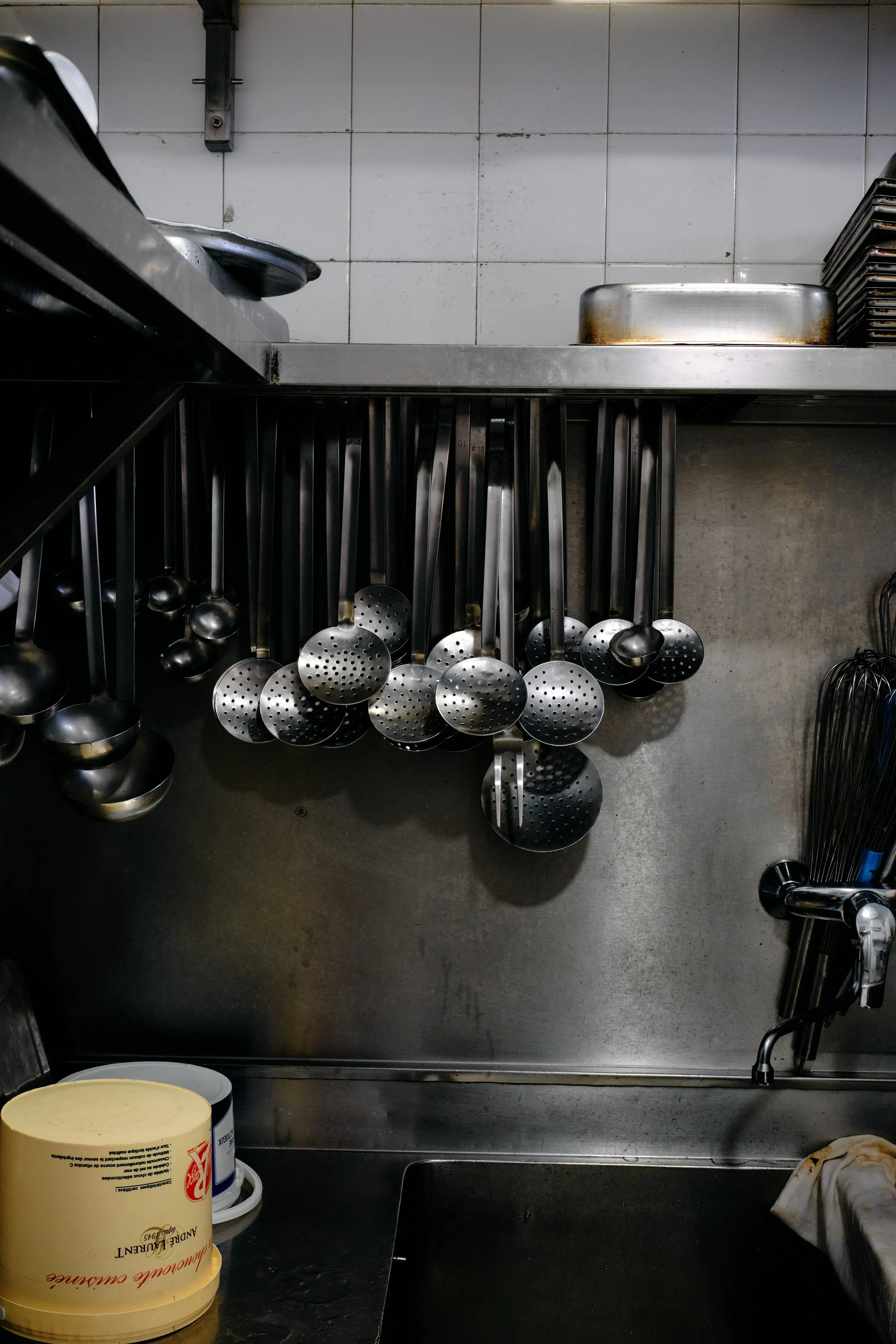 Vue de la zone de cuisine commerciale avec un évier en acier inoxydable, une étagère avec des cuillères en acier suspendues, une partie de la cuisine et des contenants en plastique, dont un contenant de glace ou de crème.
