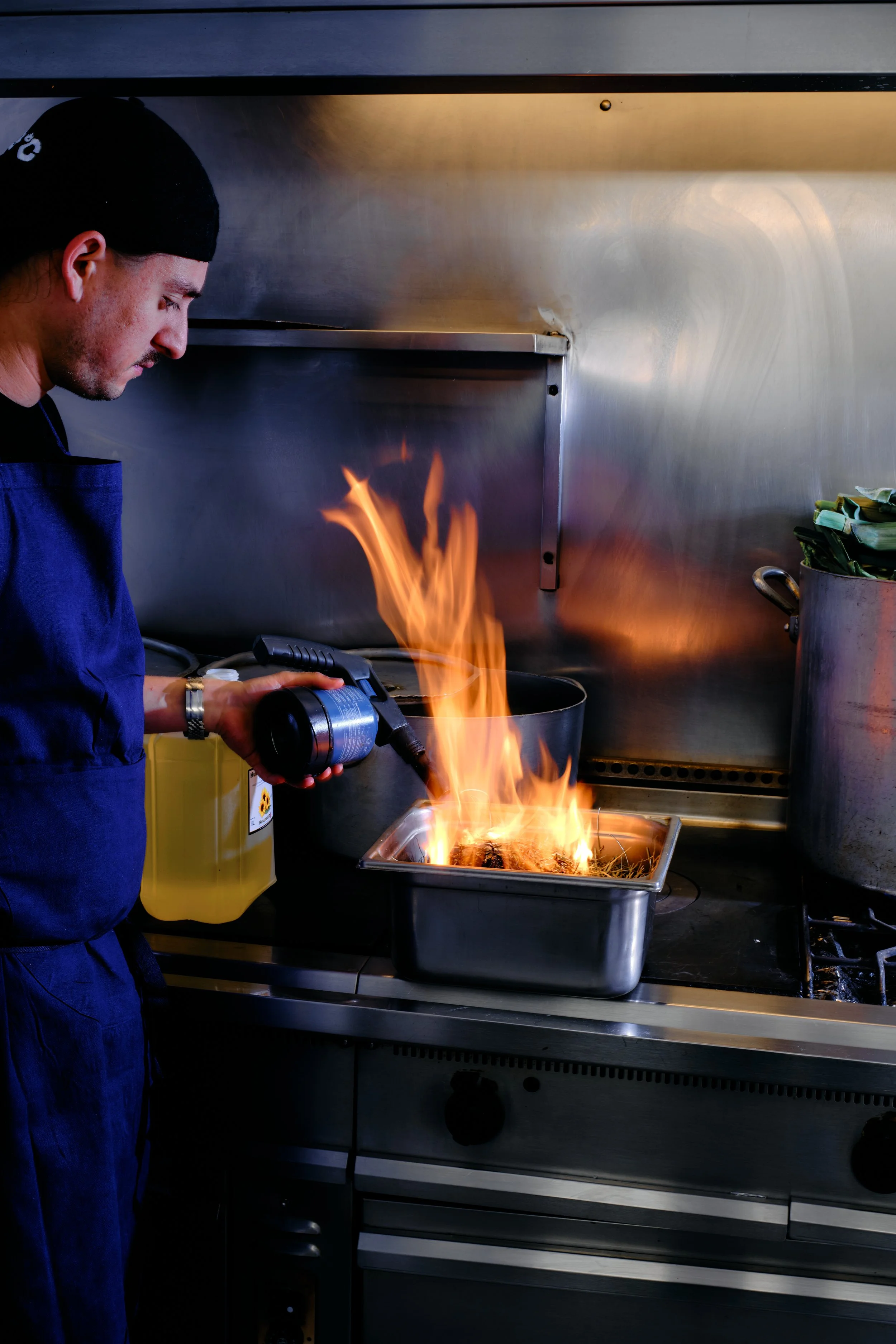Un chef en cuisine utilise un chalumeau pour flamber des aliments dans une casserole en acier inoxydable, dans une cuisine professionnelle.