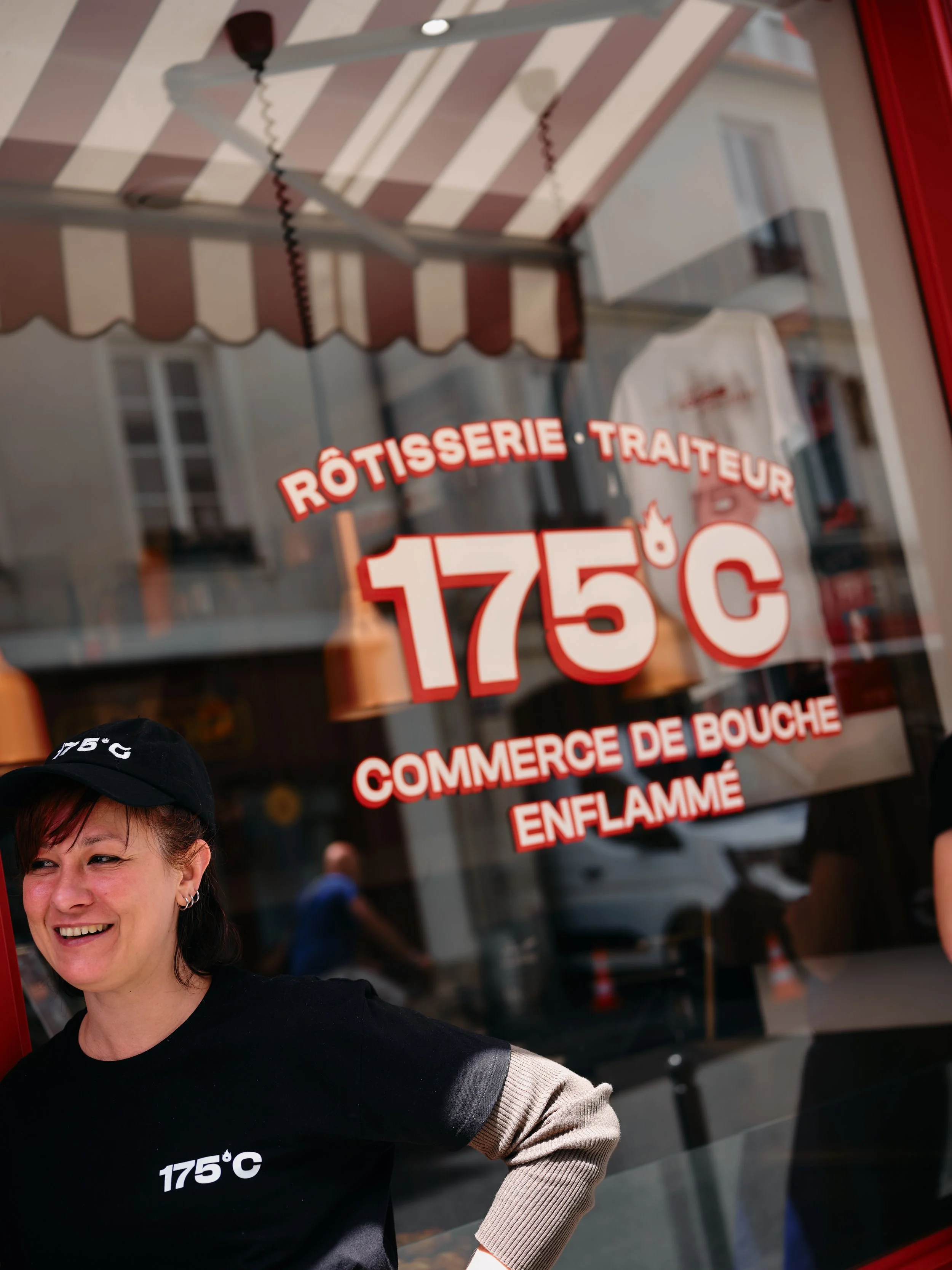 Une femme souriante portant un t-shirt noir avec la température 175°C écrit dessus, debout devant une vitrine de rôtisserie ou de magasin de viande, avec des enseignes en français indiquant la vente de viande en gros et à l'étalage.