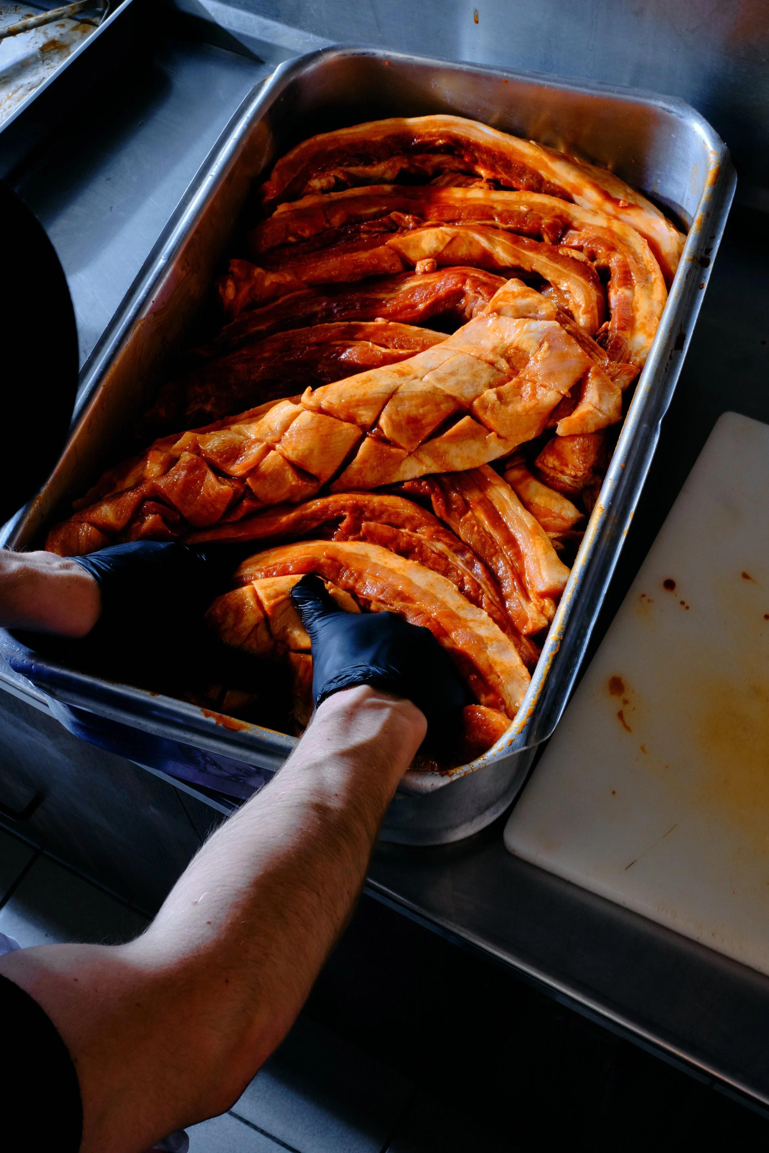 Une personne portant des gants noirs manipule de la viande de porc marinée dans un grand plat en inox. La viande est coupée en morceaux et en longues tranches, prête à être cuisinée.
