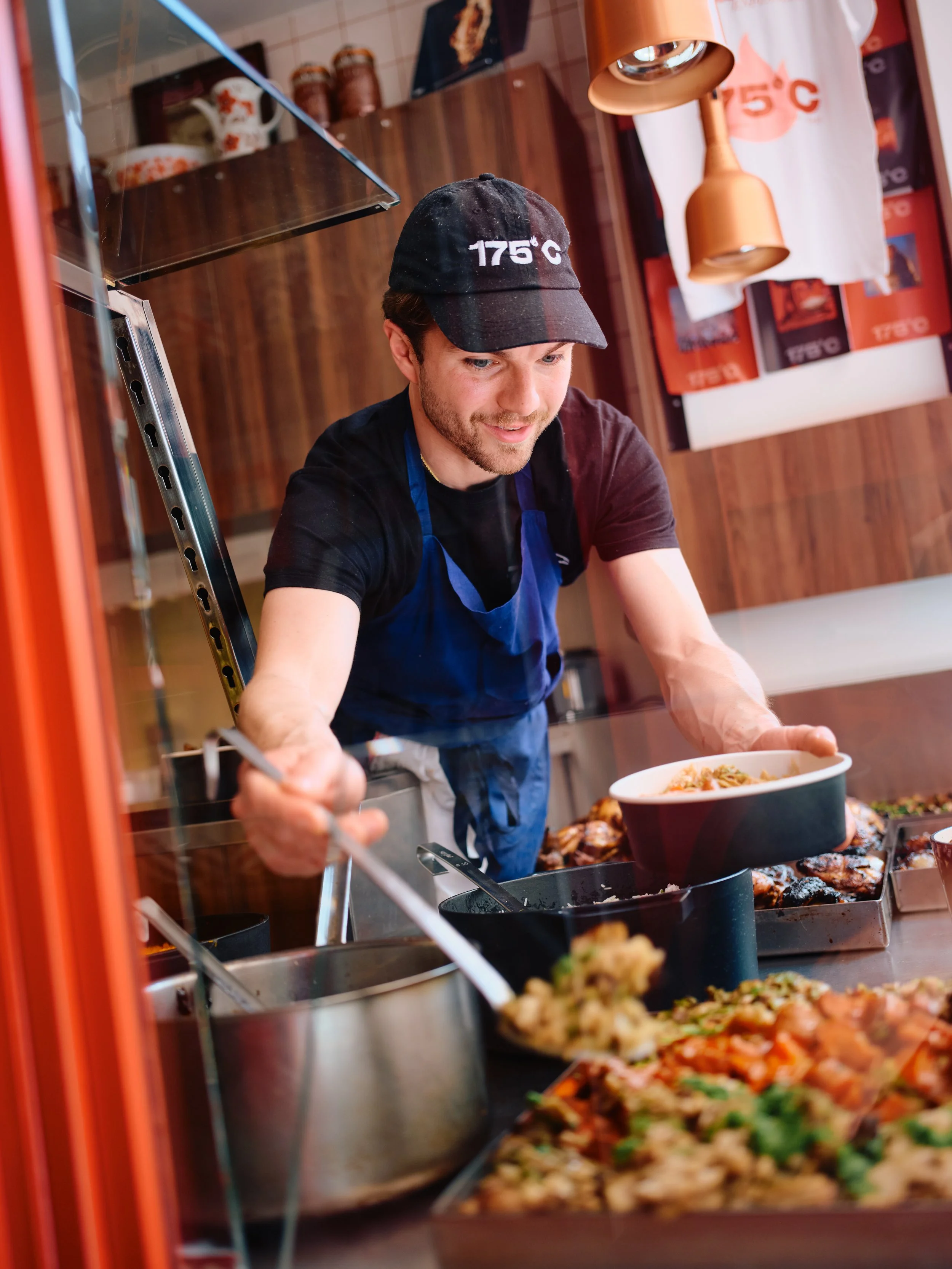 Jeune homme souriant portant une casquette noire avec la température 175 °C dessus, servant de la nourriture dans une rôtisserie de quartier du 11e arrondissement de Paris, avec plusieurs plats en préparation devant lui.