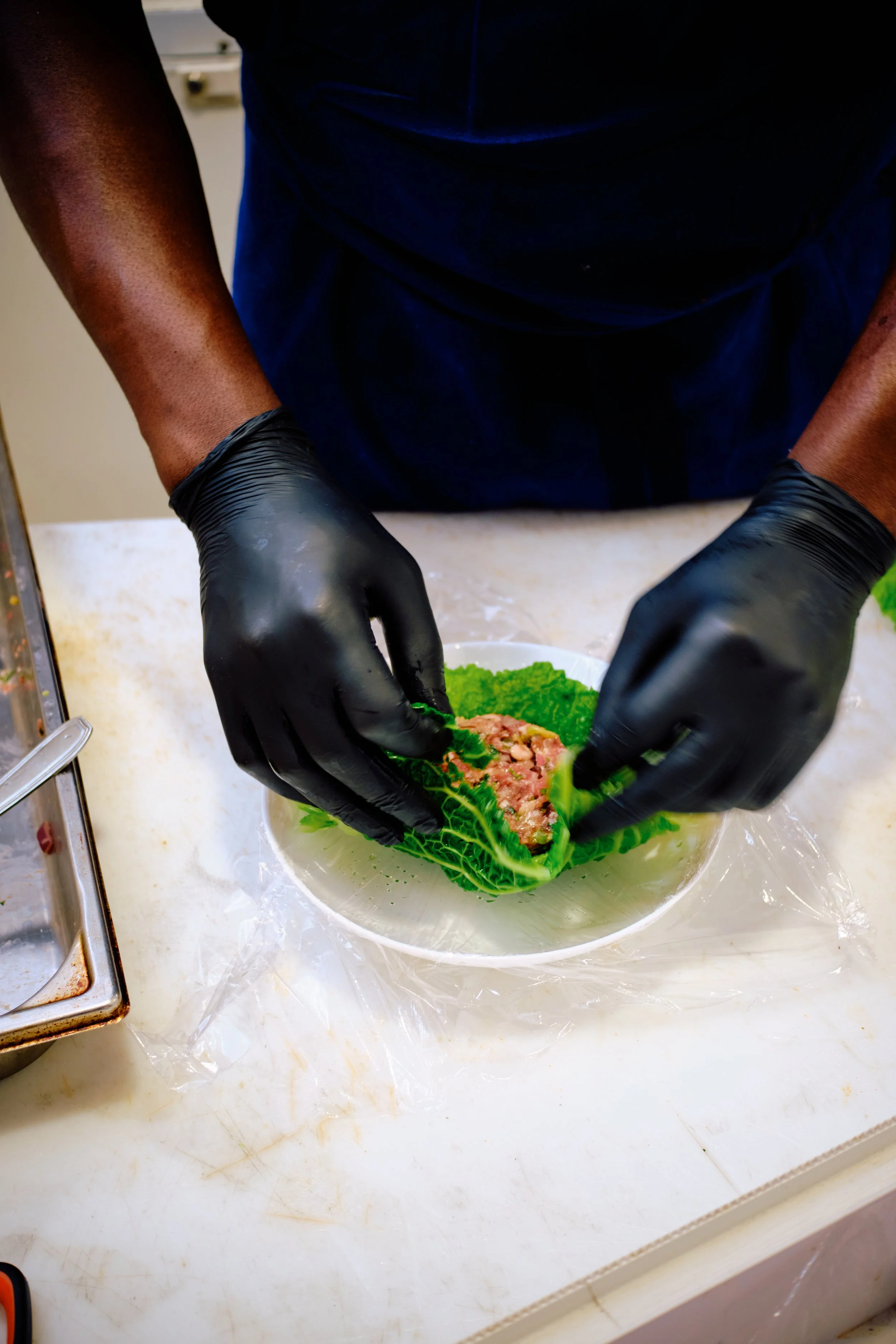 Une personne portant des gants noirs prépare de la viande hachée dans une feuille de chou verte dans un bol blanc sur une surface en plastique.
