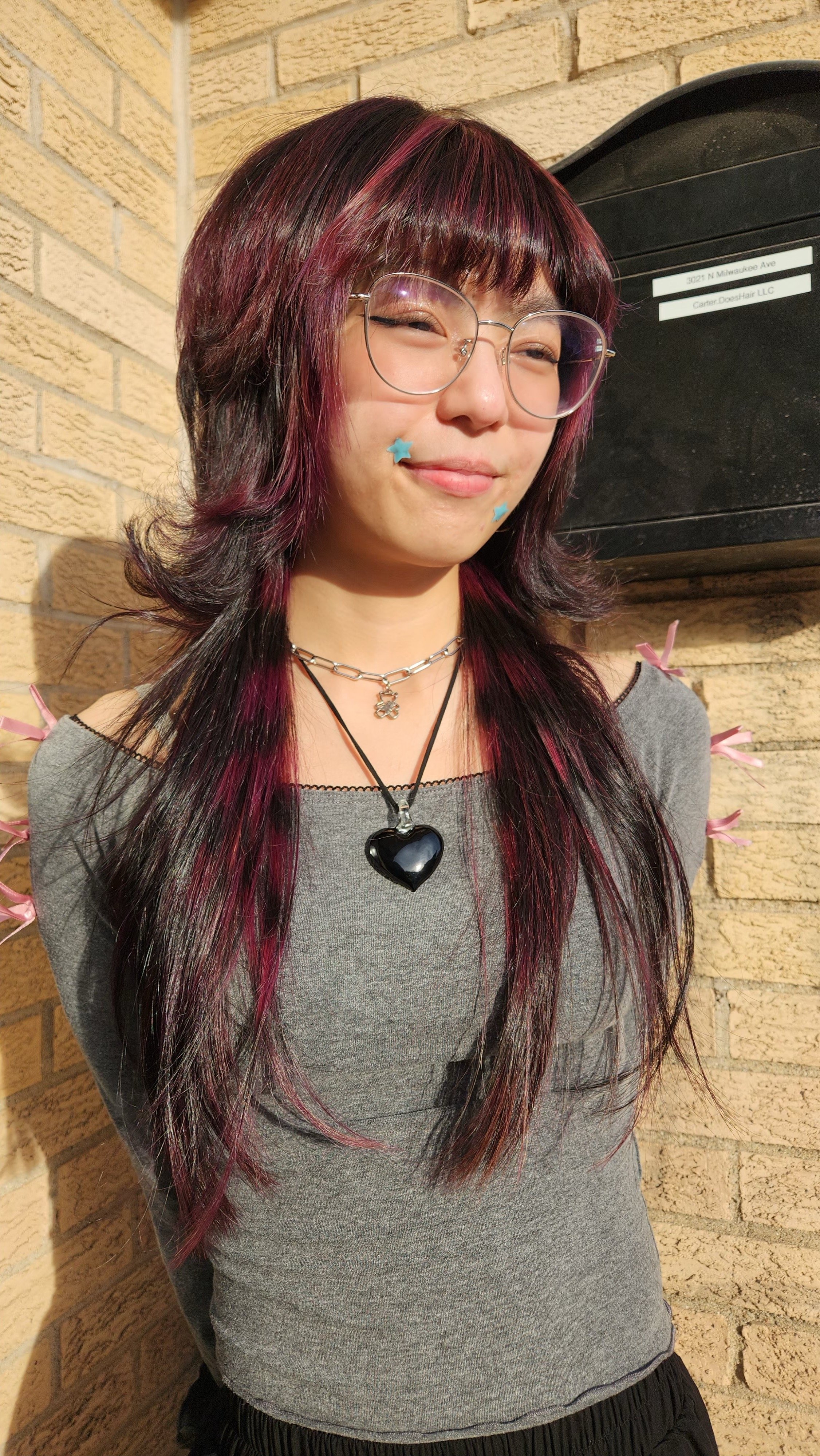 Young woman with multicolored hair, wearing glasses, a grey top, and layered necklaces, standing against a brick wall with sunlight shining on her face.
