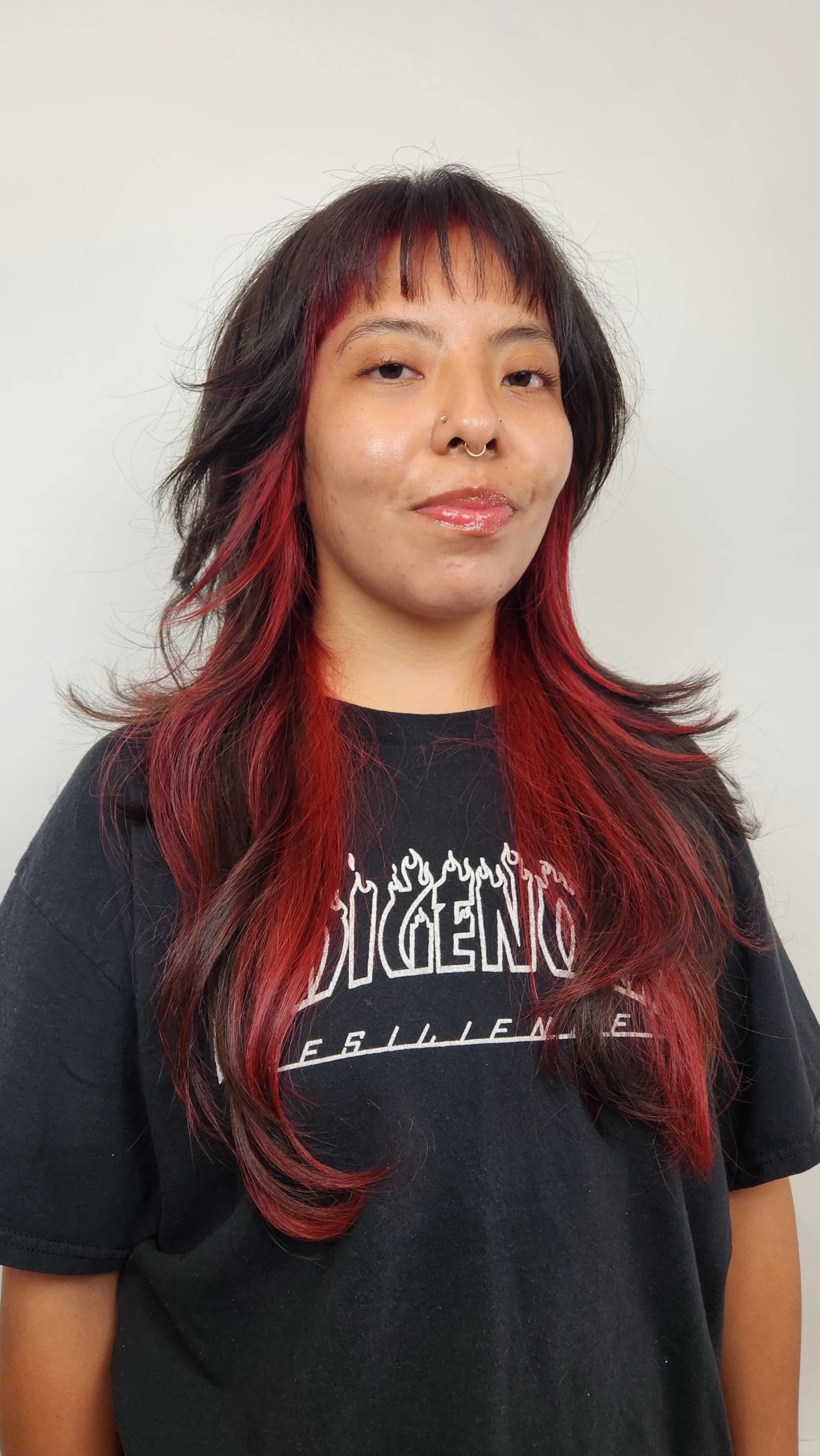 A woman with layered black and red hair, wearing a black t-shirt with graphic text. She has a nose piercing and is standing against a plain light-colored wall.
