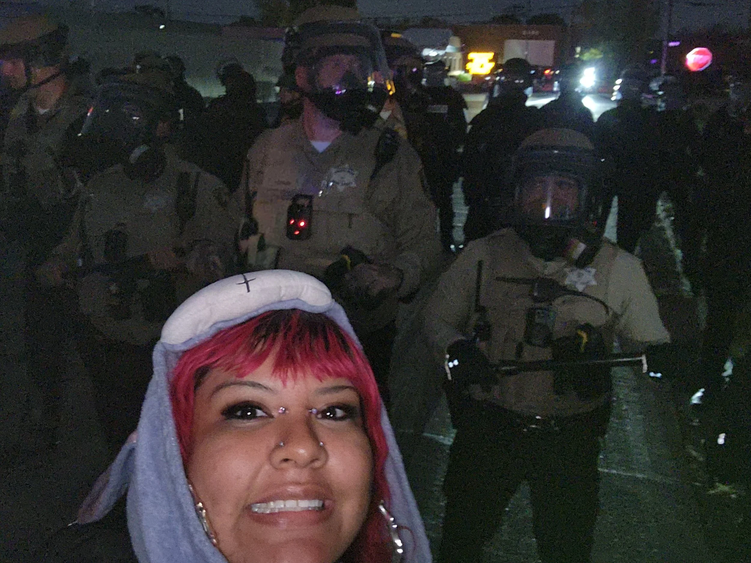 A woman with pink hair and piercings taking a selfie in front of a group of police officers wearing riot gear at night.