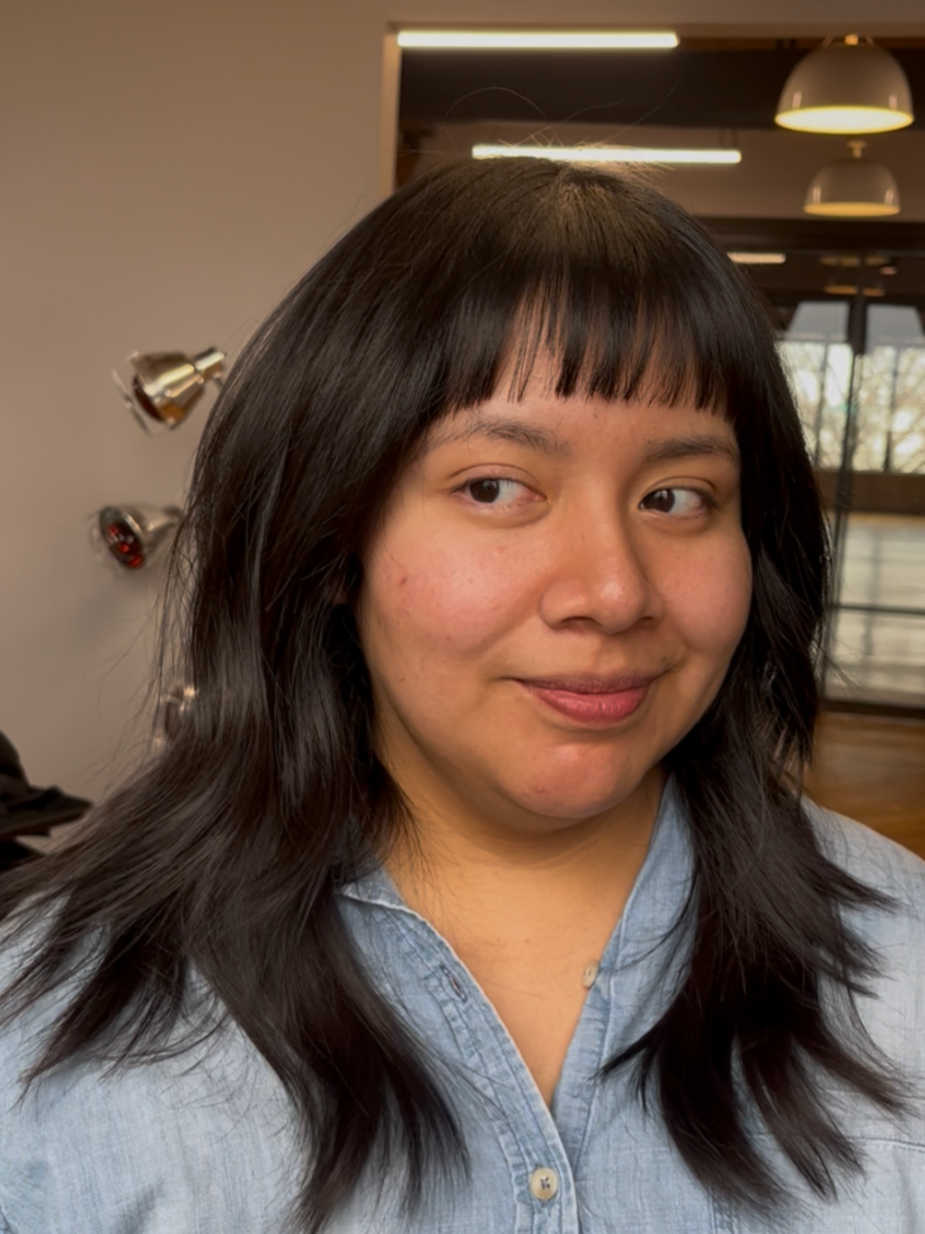 A woman with shoulder-length dark hair and bangs, wearing a light blue denim shirt, standing indoors with warm lighting and large windows in the background.