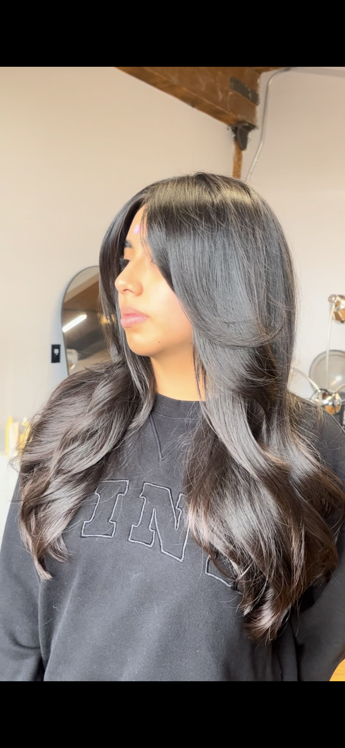 A woman with long, wavy black hair with subtle highlights is looking to the side in a salon or studio setting.