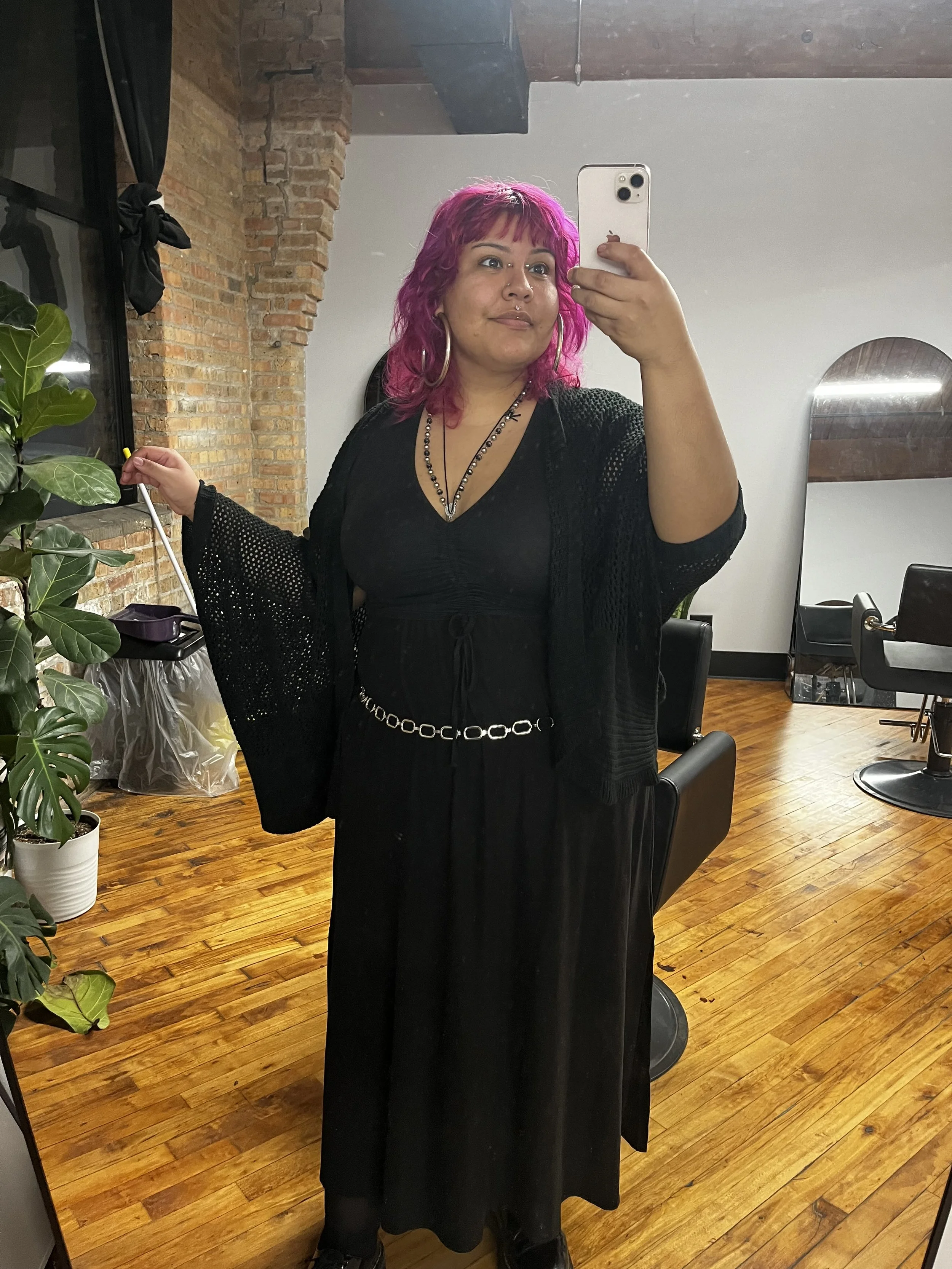 Woman with pink hair wearing a black outfit, standing in a salon, taking a selfie in a mirror.