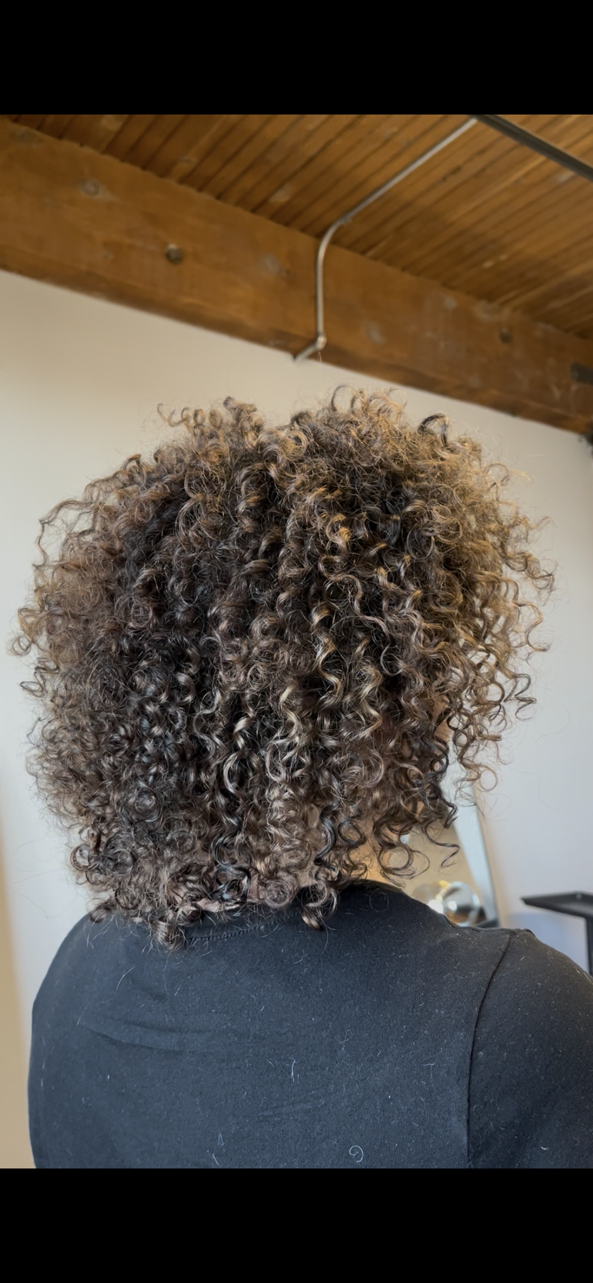 Back view of a person with short, tightly curled, brown hair. They are wearing a black shirt and are indoors with a wooden ceiling and a white wall in the background.
