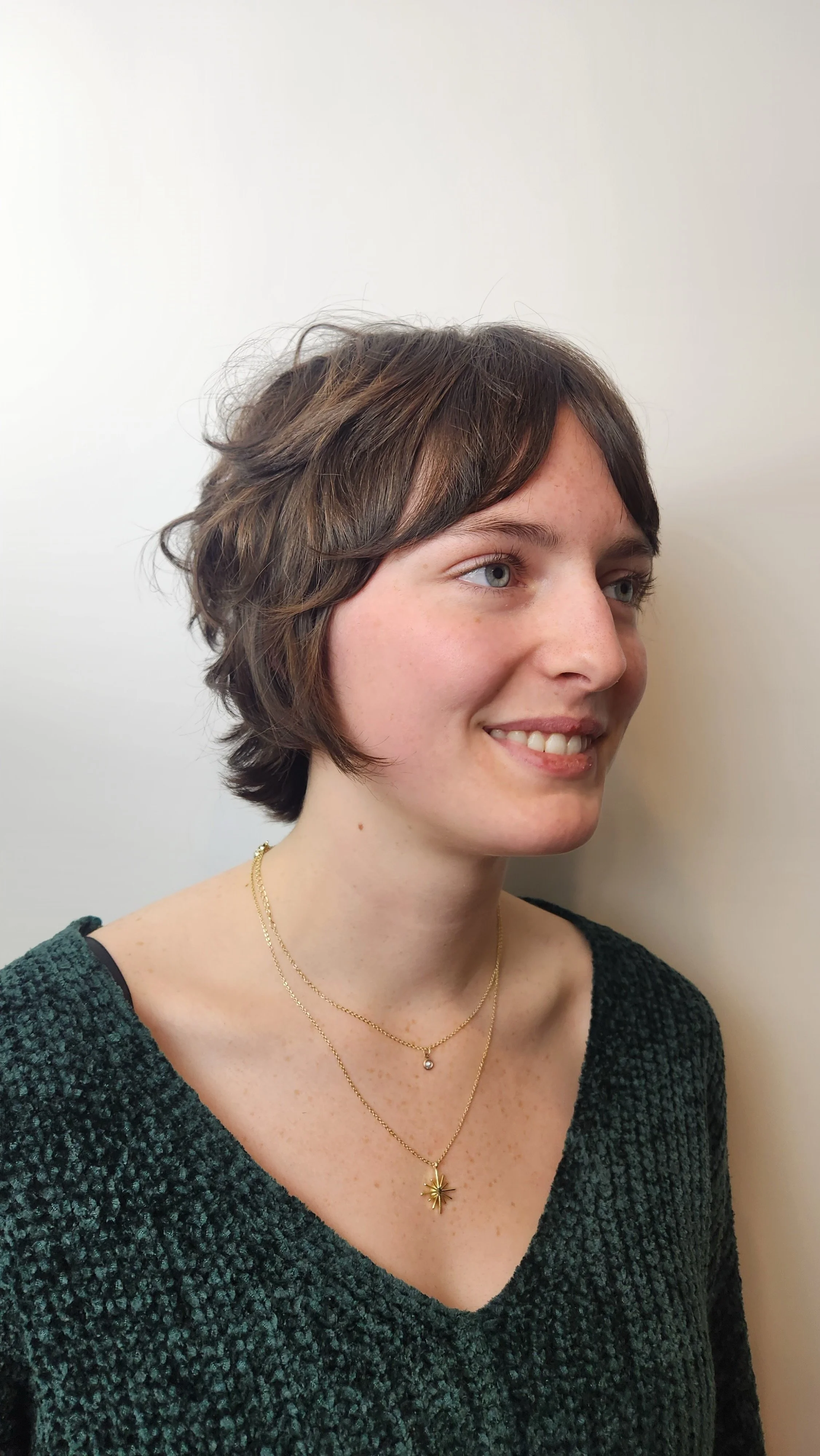 A young woman with short brown hair and blue eyes wearing layered gold necklaces and a dark green sweater, smiling and looking to the right, standing against a plain white wall.