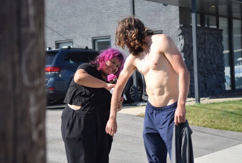 A woman with pink hair is smiling and speaking into a microphone held by a shirtless young man with long brown hair, standing outdoors near a modern building.