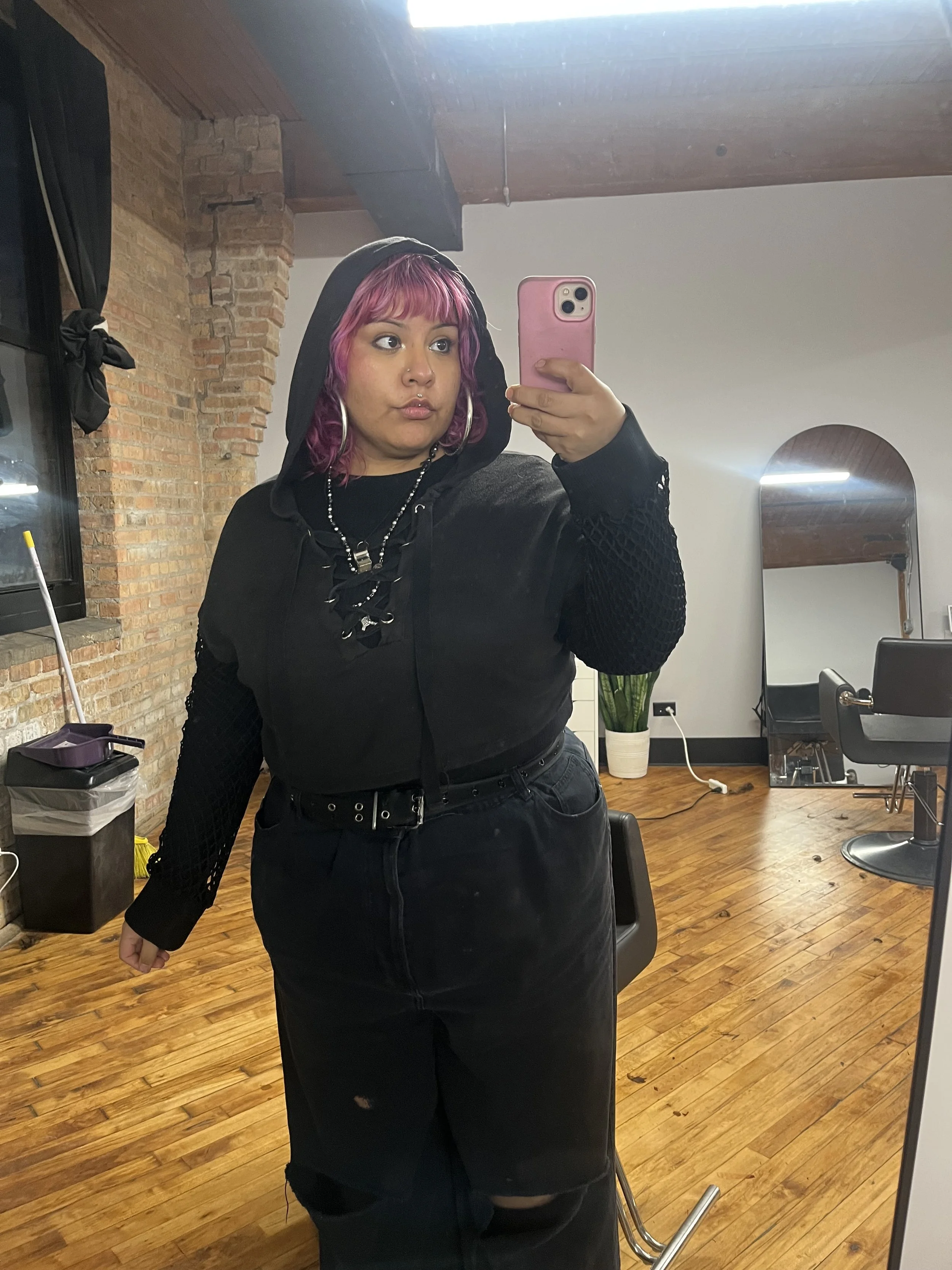 Woman with pink and purple hair taking a selfie in a mirror, wearing a black hoodie under a black jacket, and black pants with a small rip.