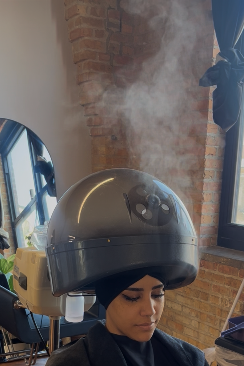 Woman sitting under a hairdryer at a salon, with steam rising from the dryer, brick wall and window in the background.
